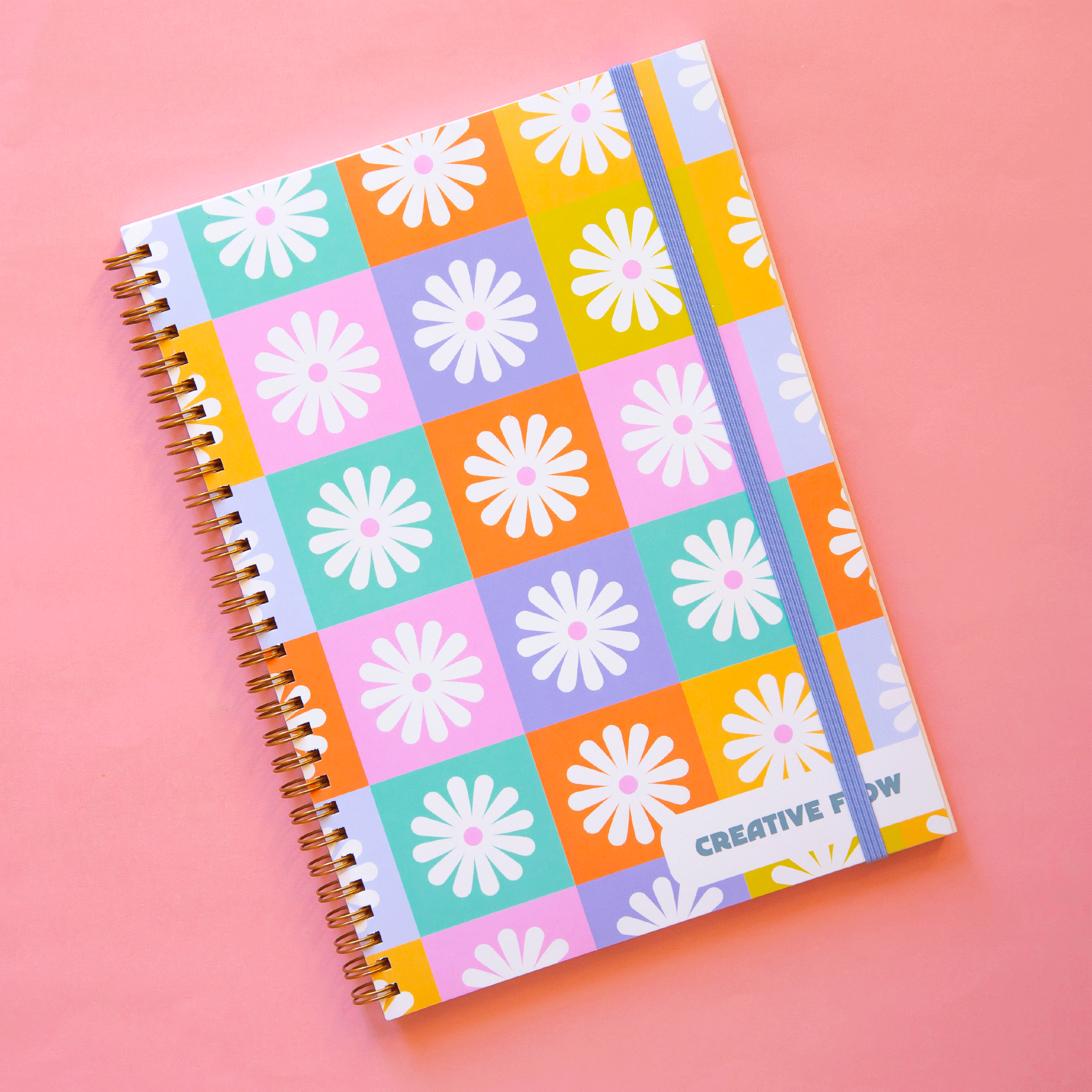  A spiral bound notebook with a multicolored checker design that has a white daisy in the center of each square. In the bottom right hand corner it says, "Creative Flow" in green text.