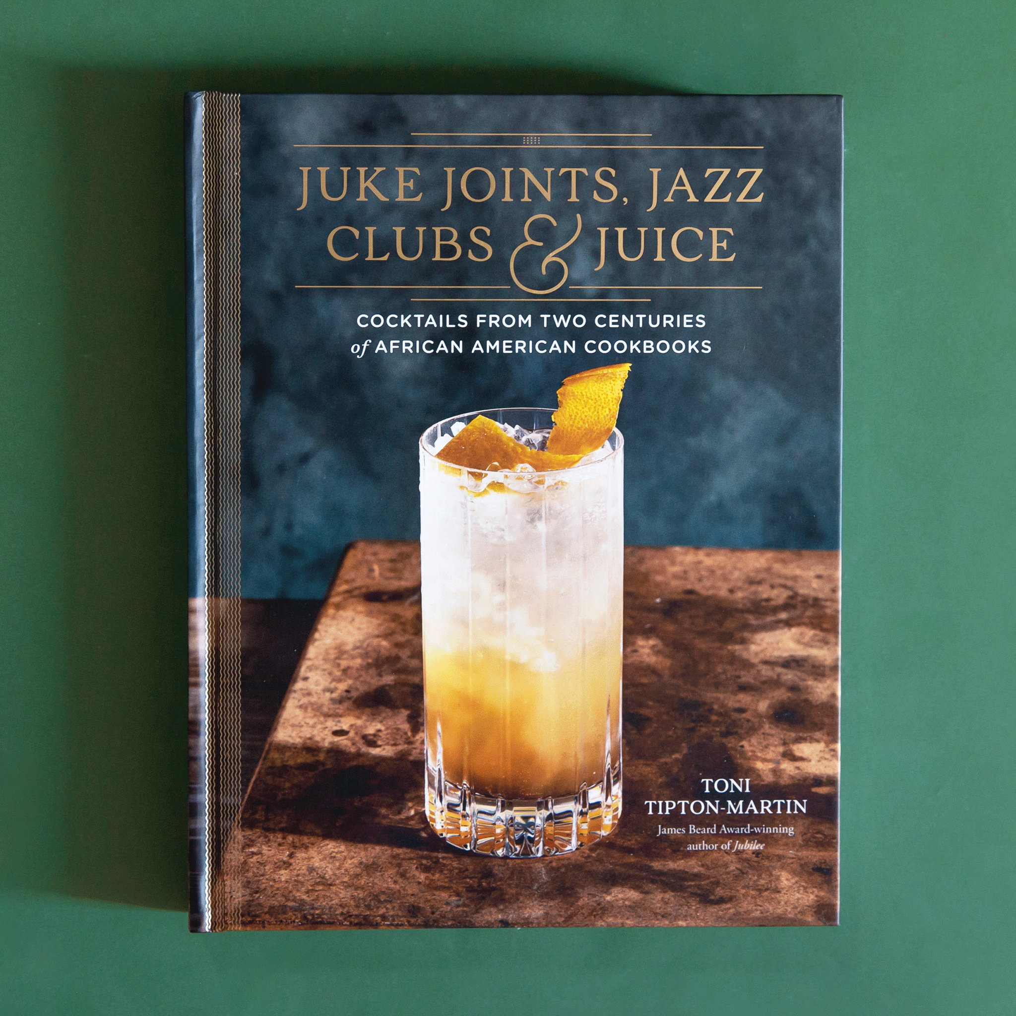 On a green background is a grey and brown book cover with a yellow cocktail and gold text that reads, &quot;Juke Joints, Jazz Clubs &amp; Juice&quot;, &quot;Cocktails From Two Centuries of African American Cookbooks&quot;.