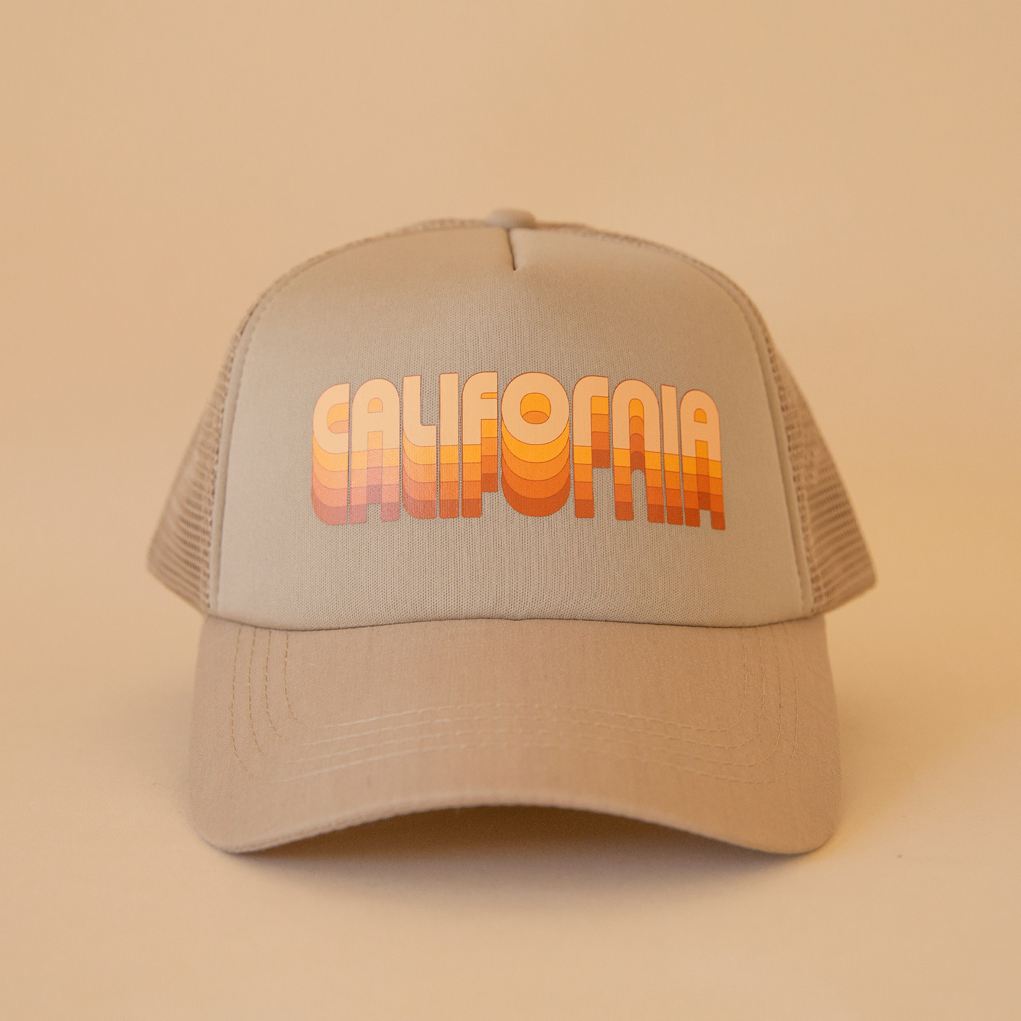 On a tan background is a tan trucker hat with yellow and orange gradient text that reads, &quot;California&quot;.