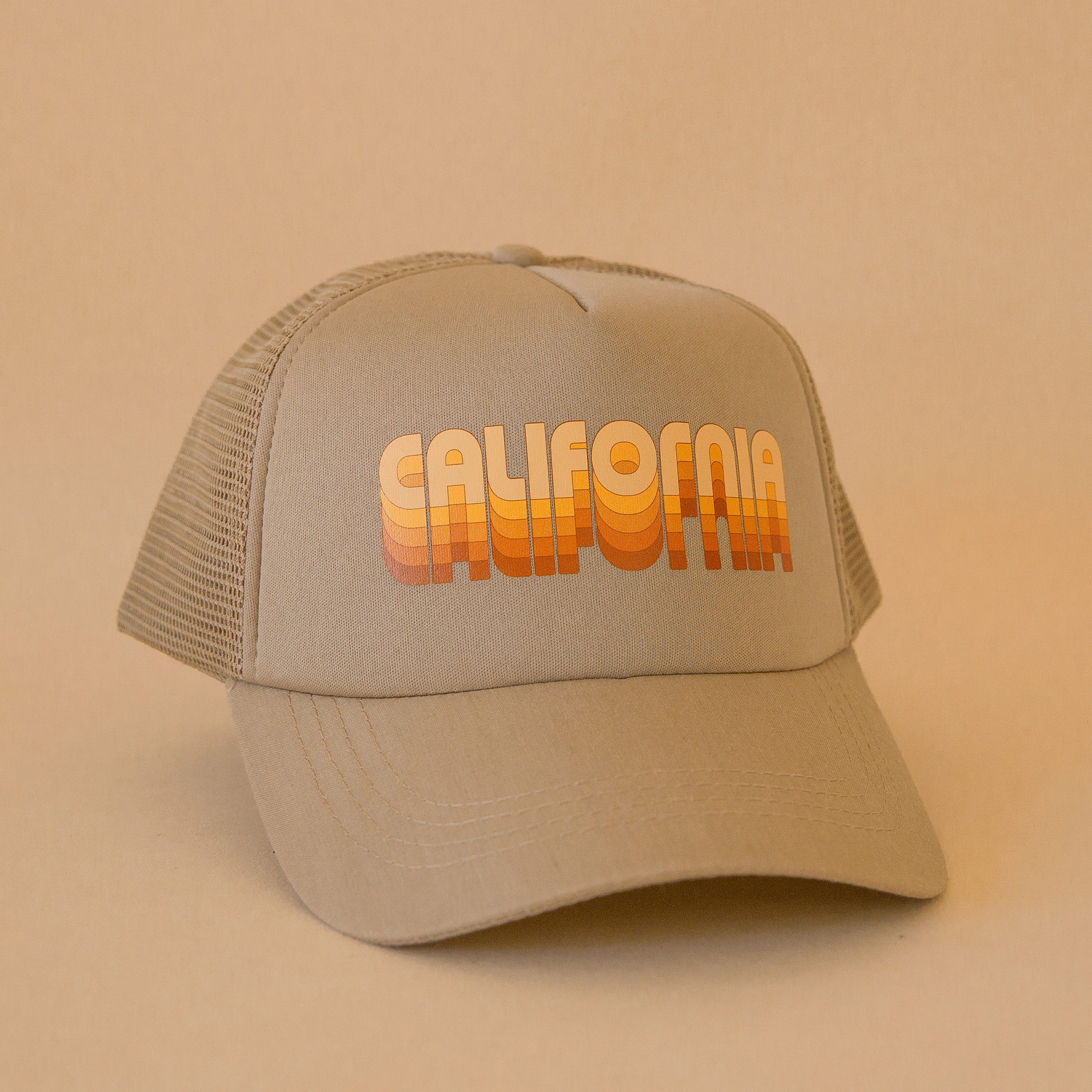 On a tan background is a tan trucker hat with yellow and orange gradient text that reads, &quot;California&quot;.