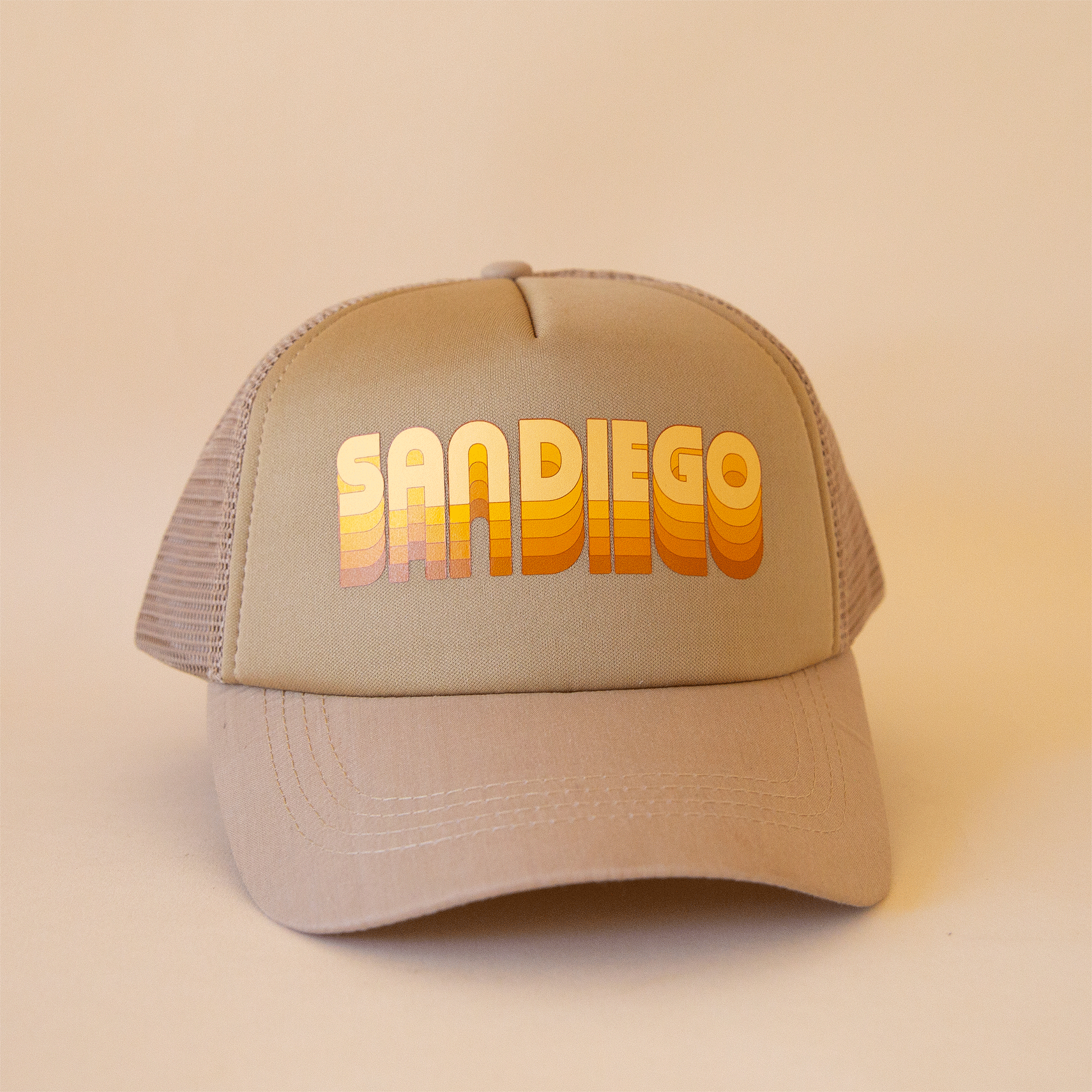 On a tan background is a tan trucker hat with yellow and orange gradient text that reads, "San Diego".