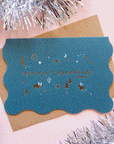 On a light pink background is a teal blue wavy edged greeting card with gold text and details that read, "season's greetings" along with a Kraft brown envelope. 