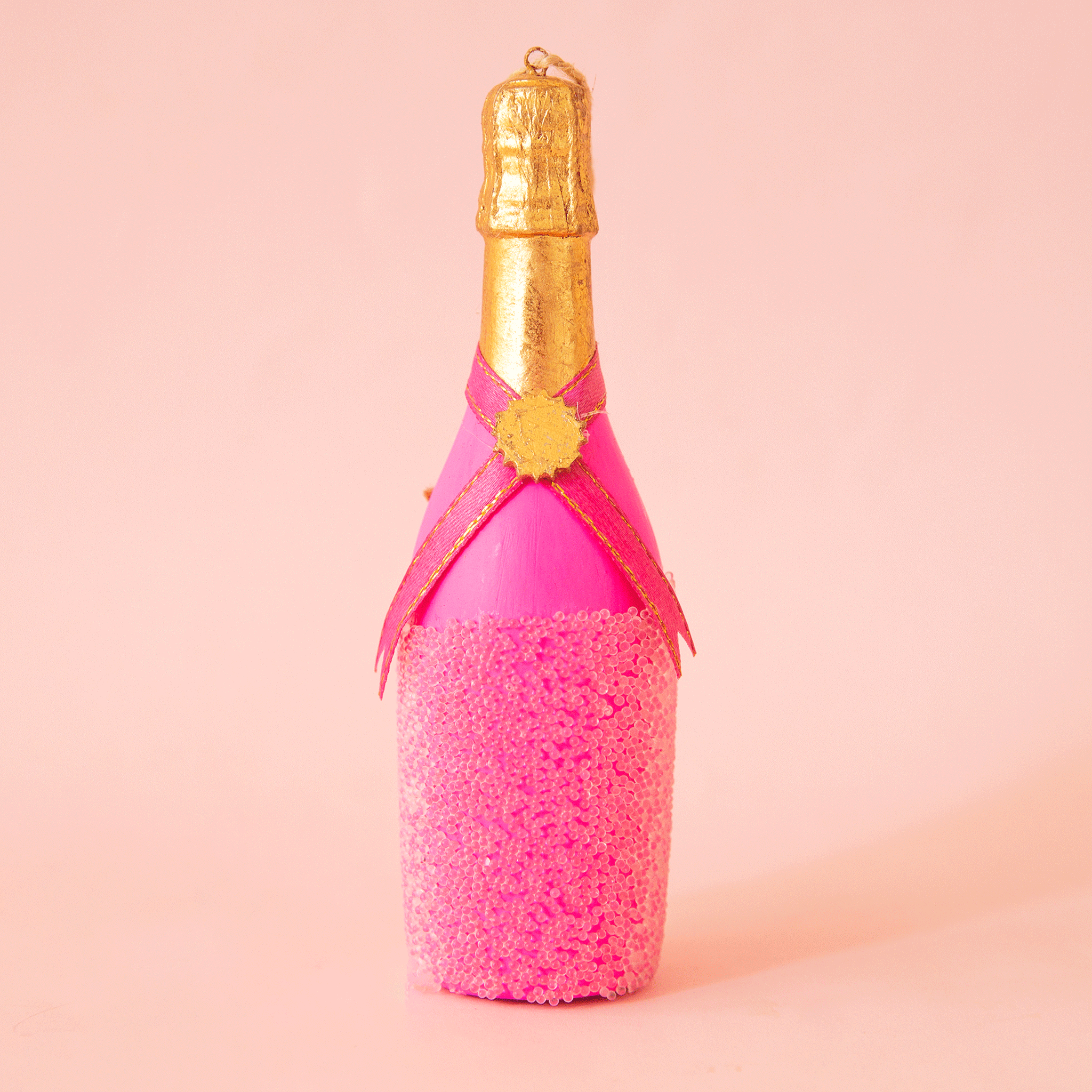 On a peach background is a resin champagne bottle shaped ornament with a pink bottle and a gold top.