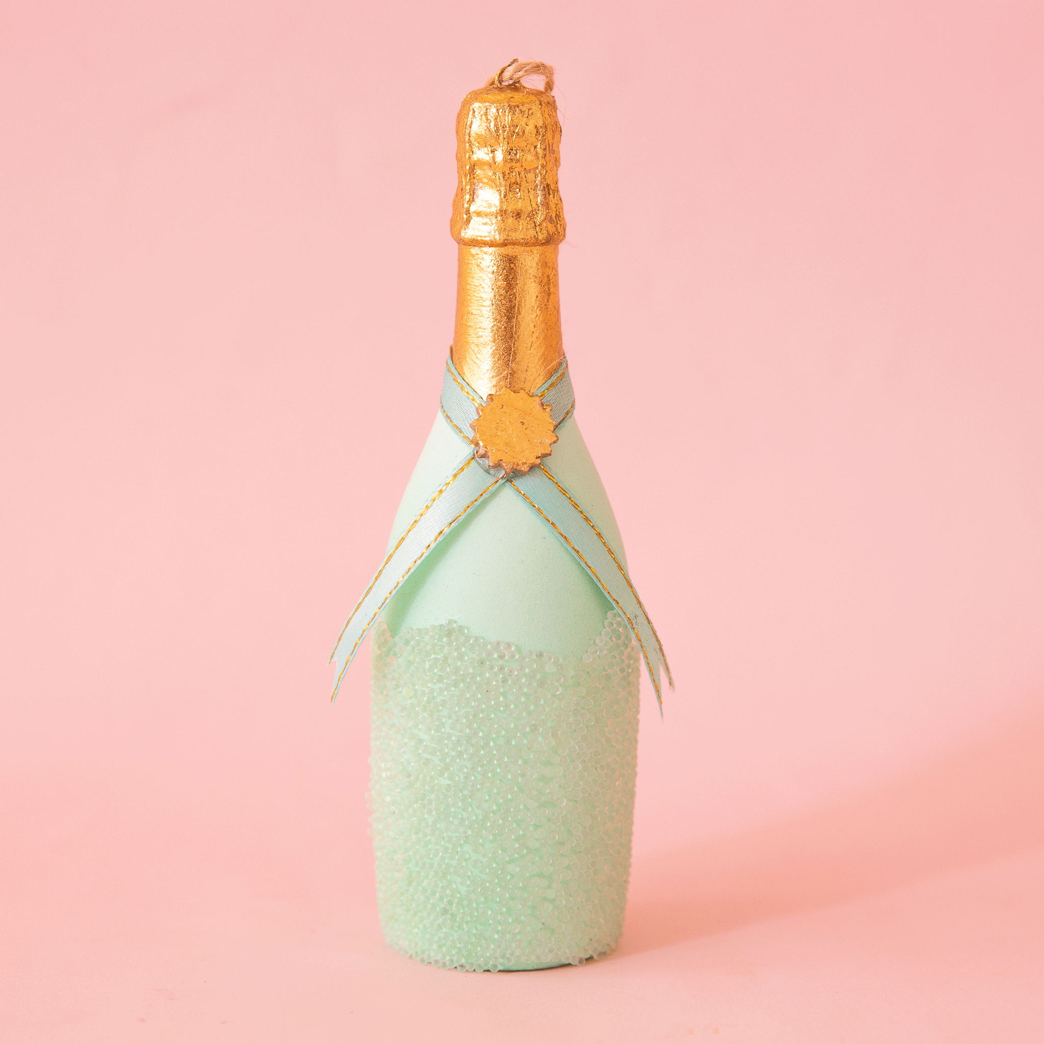 On a pink background is a mint green champagne bottle shaped ornament.