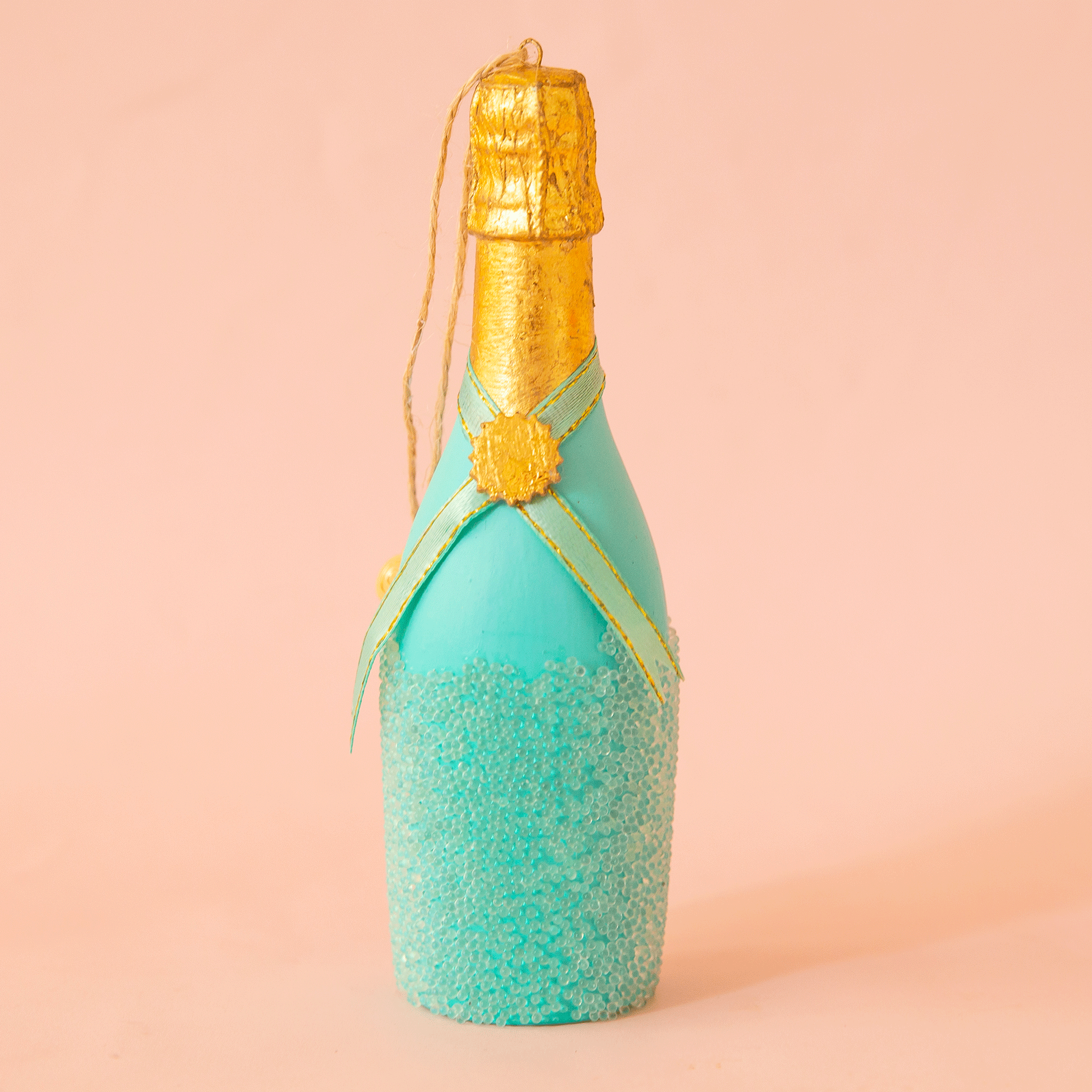 On a pink background is a turquoise champagne bottle shaped ornament. 
