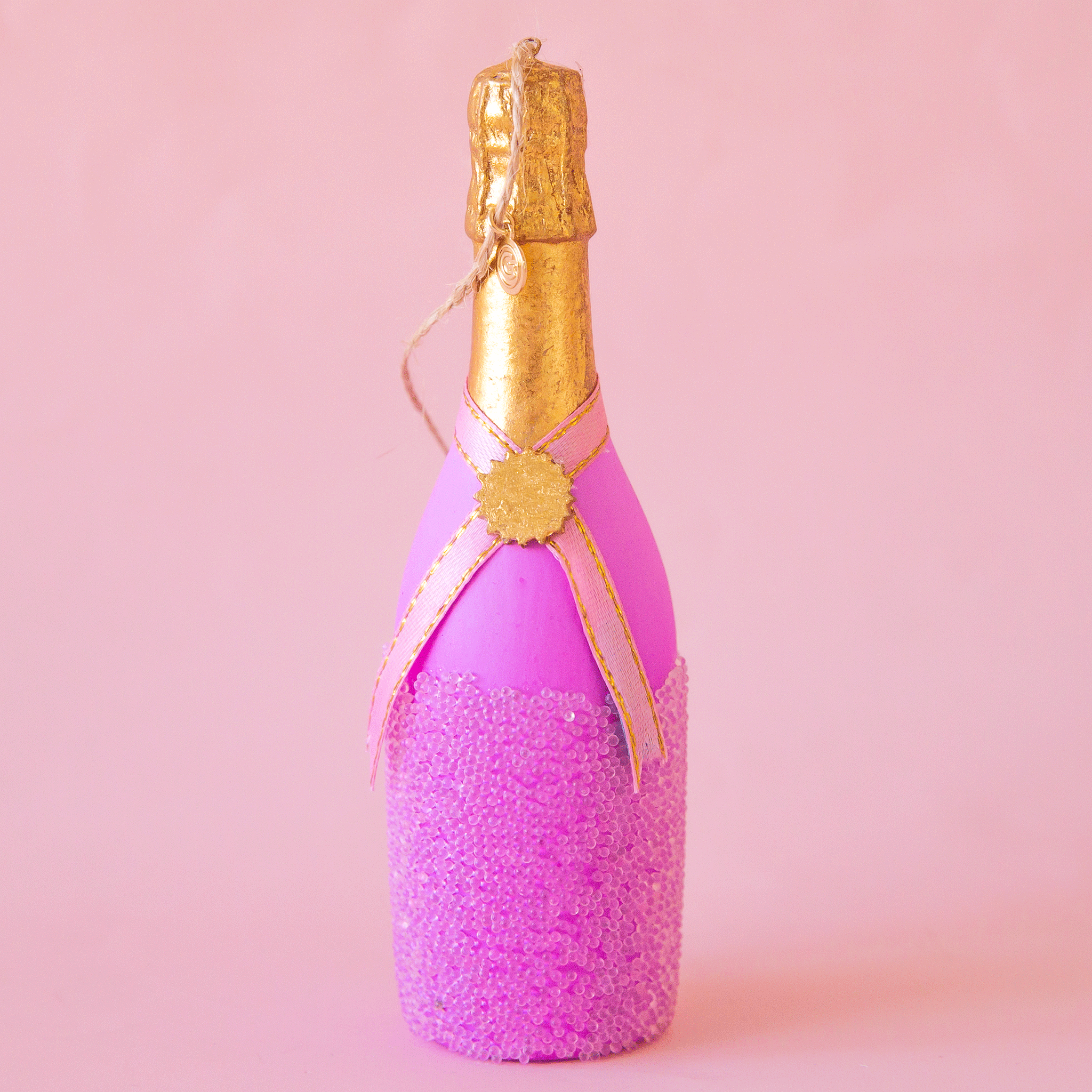 On a pink background is a lilac champagne bottle shaped ornament. 
