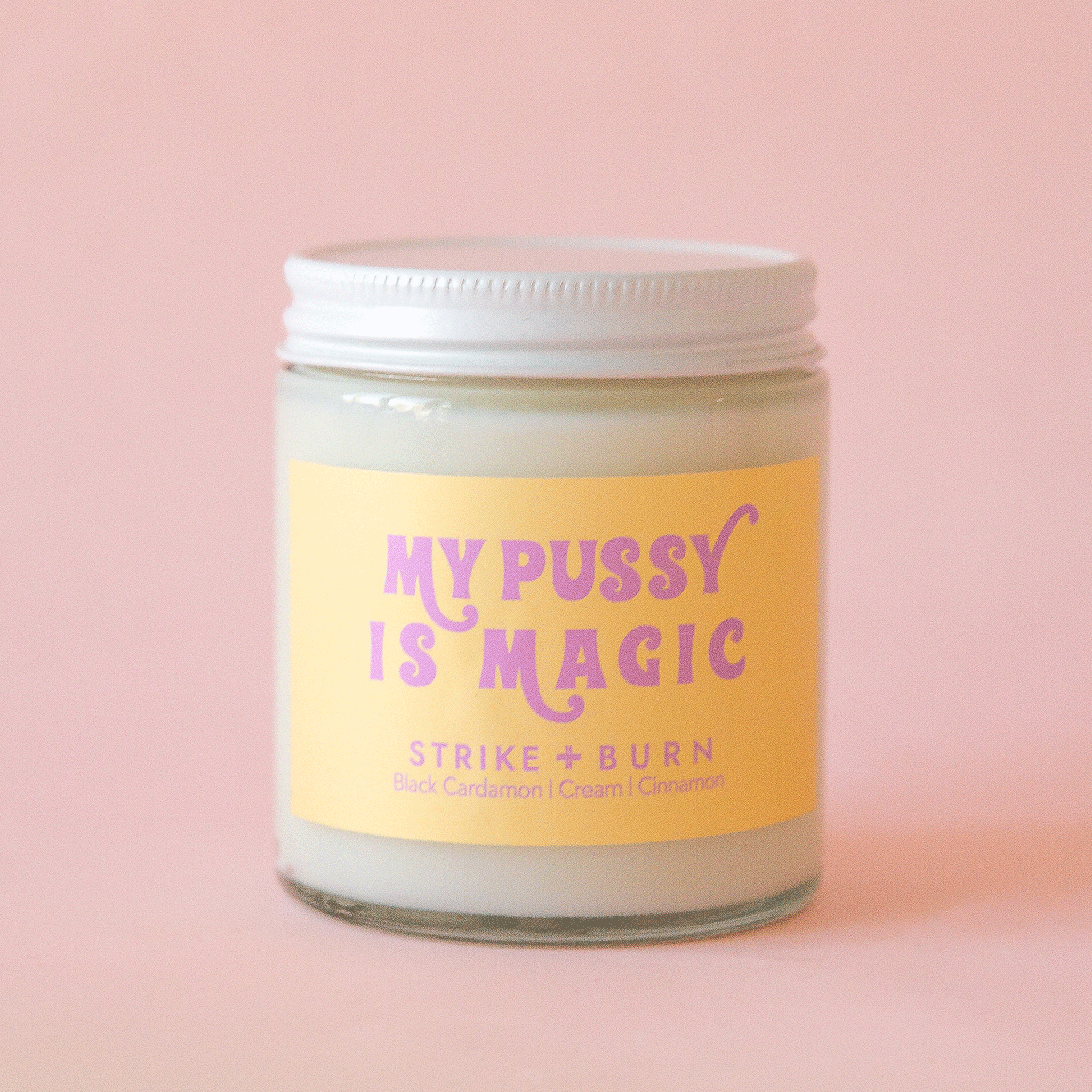 On a pink background is a white glass candle with a white lid and a yellow label on the front with pink text that reads, &quot;My Pussy Is Magic Strike + Burn&quot;.