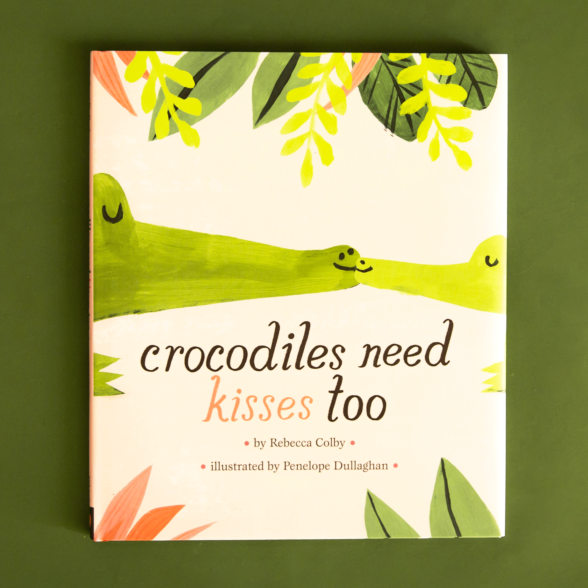 On a green background is a ivory book cover with green crocodiles kissing and black text that reads, "crocodiles need kisses too". 