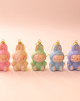 Five fuzzy pastel monster ornaments with closed eyes and gentle smiles sit in a row on a light pink background. Each has a gold hanging loop, and the colors from left to right are pink, peach, mint green, light blue, and lavender.
