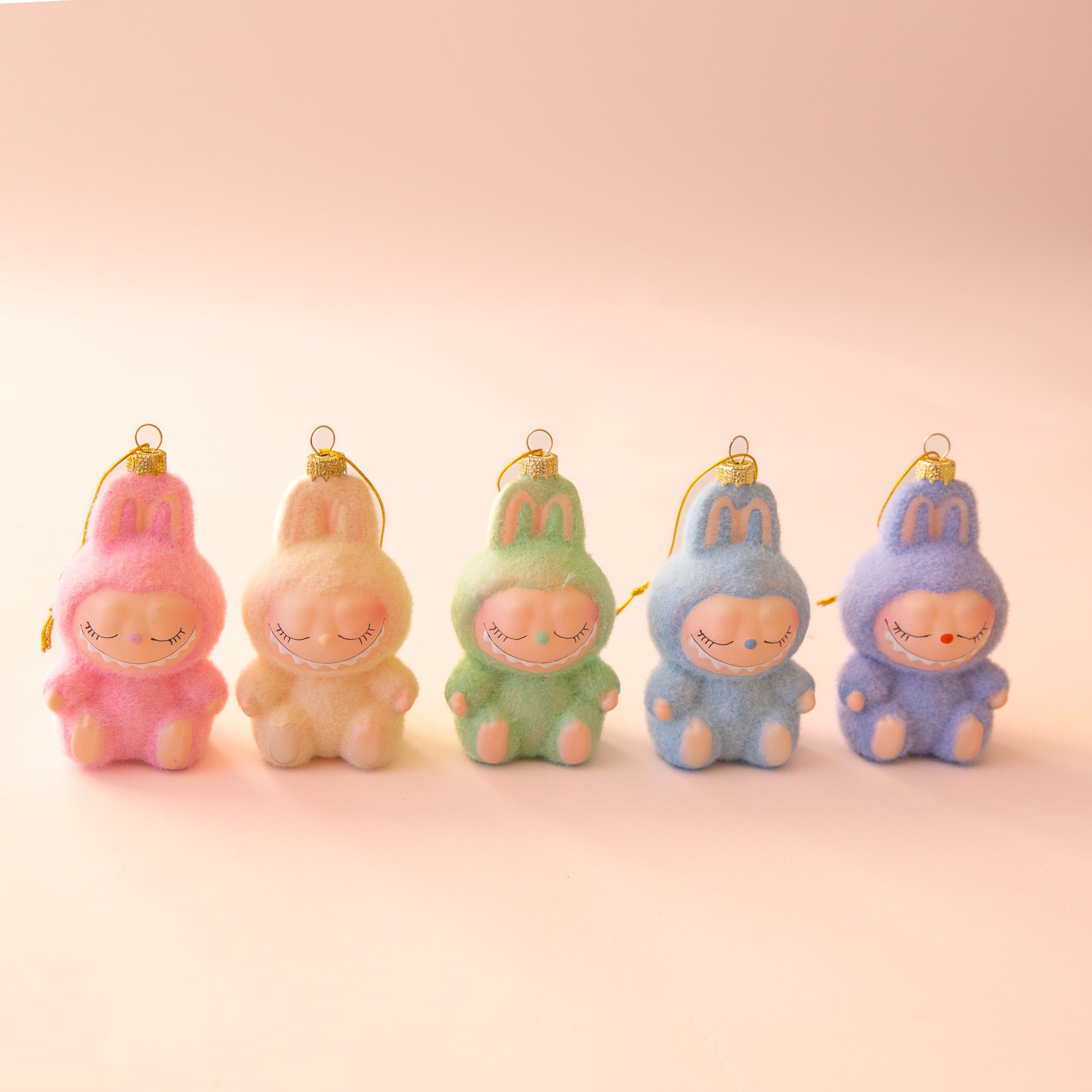 Five fuzzy pastel monster ornaments with closed eyes and gentle smiles sit in a row on a light pink background. Each has a gold hanging loop, and the colors from left to right are pink, peach, mint green, light blue, and lavender.