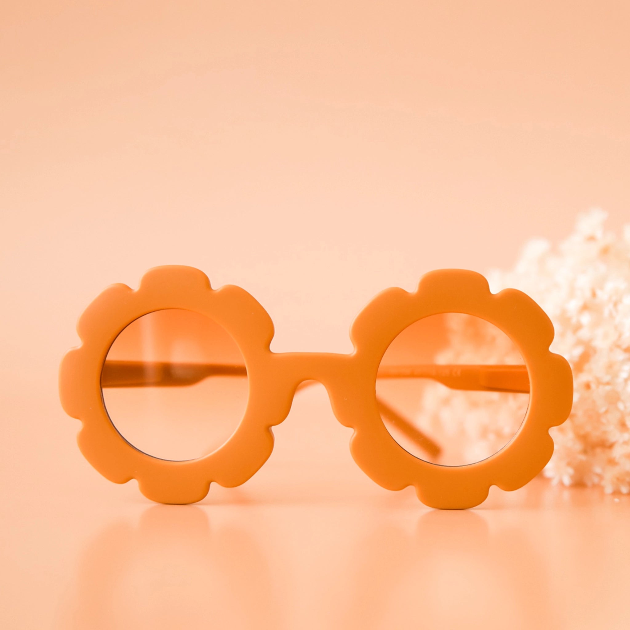 On a peachy background is an orange pair of flower shaped sunglasses with a light orange lens.