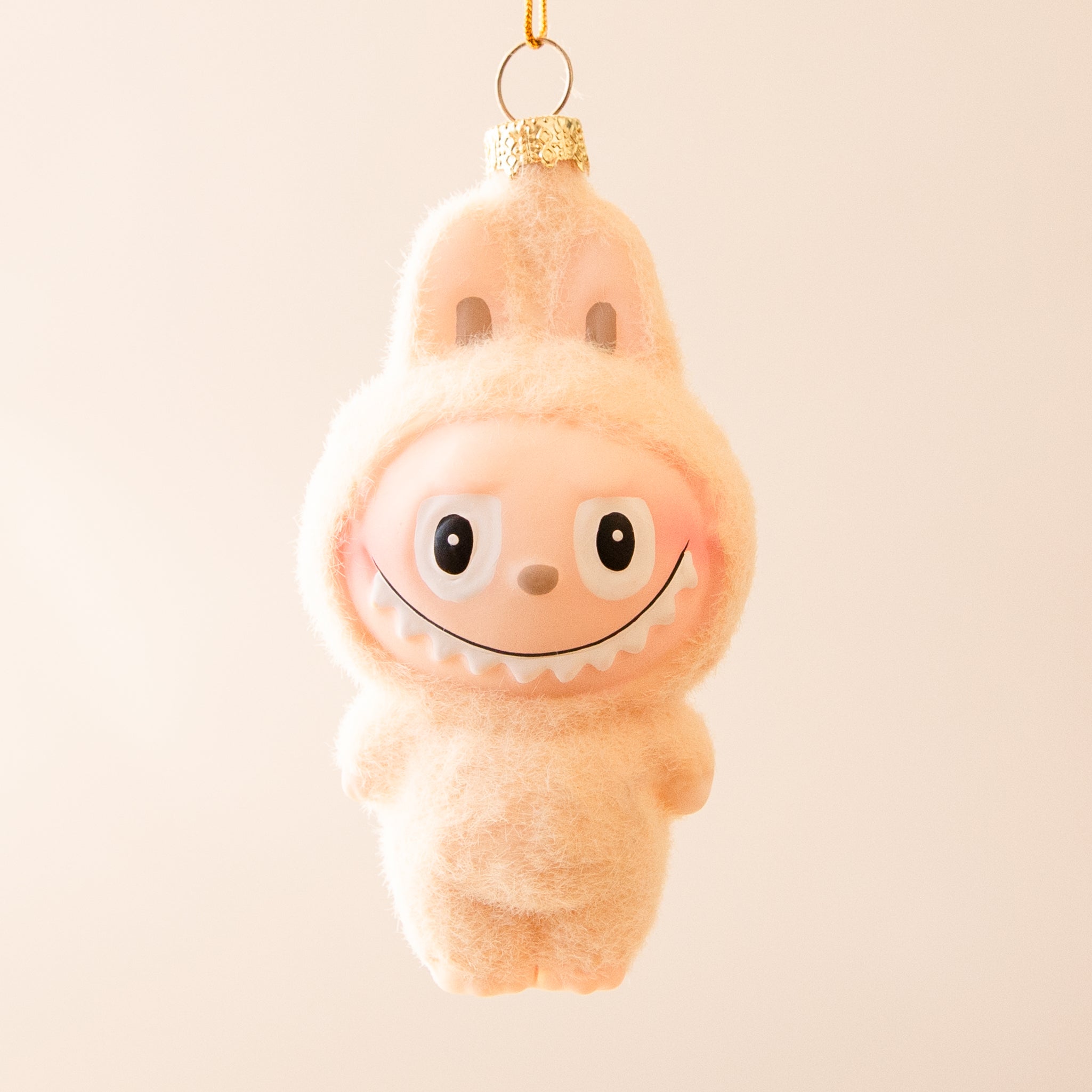 A small ivory fuzzy monster ornament with bunny-like ears and a wide toothy smile hangs from a gold string against a soft ivory background.
