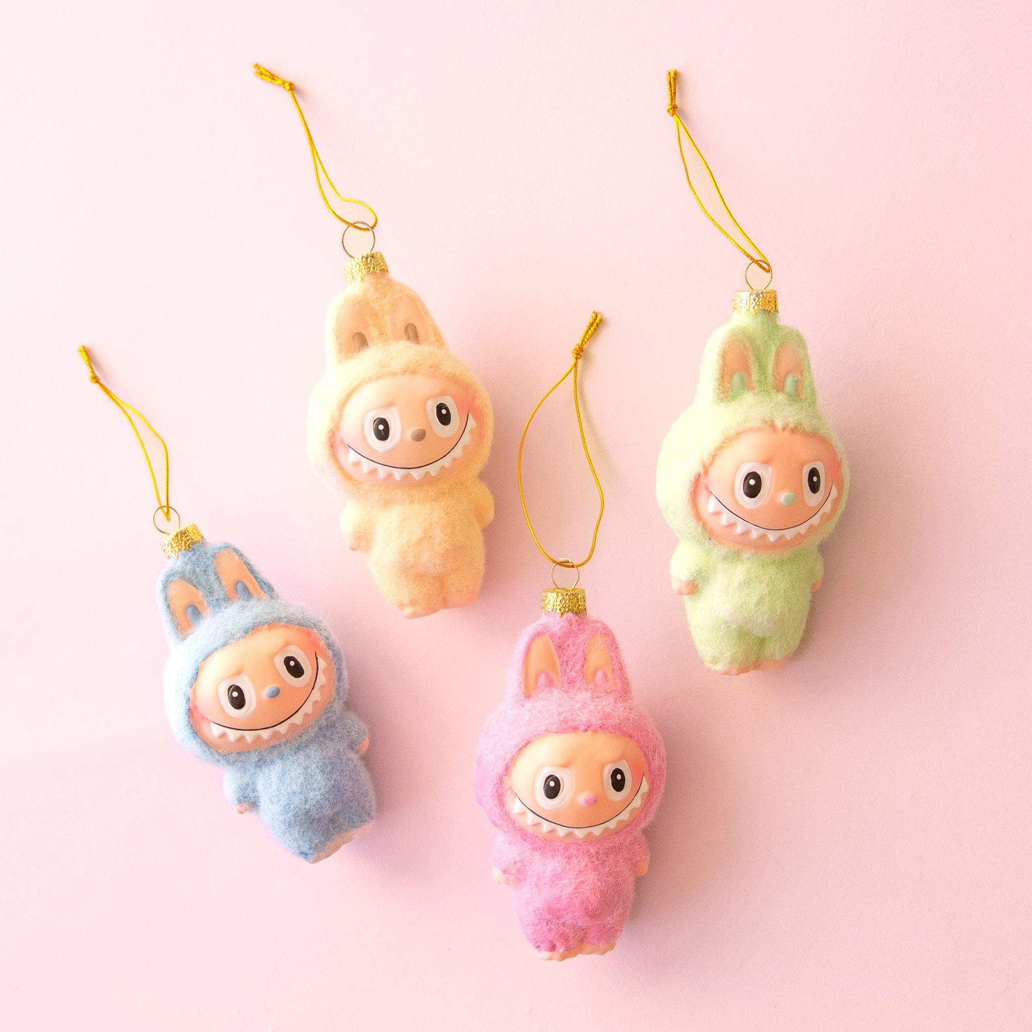 Four plush monster ornaments in pastel colors (blue, peach, pink, and green) with wide smiling faces and gold hanging strings, arranged on a light pink background.