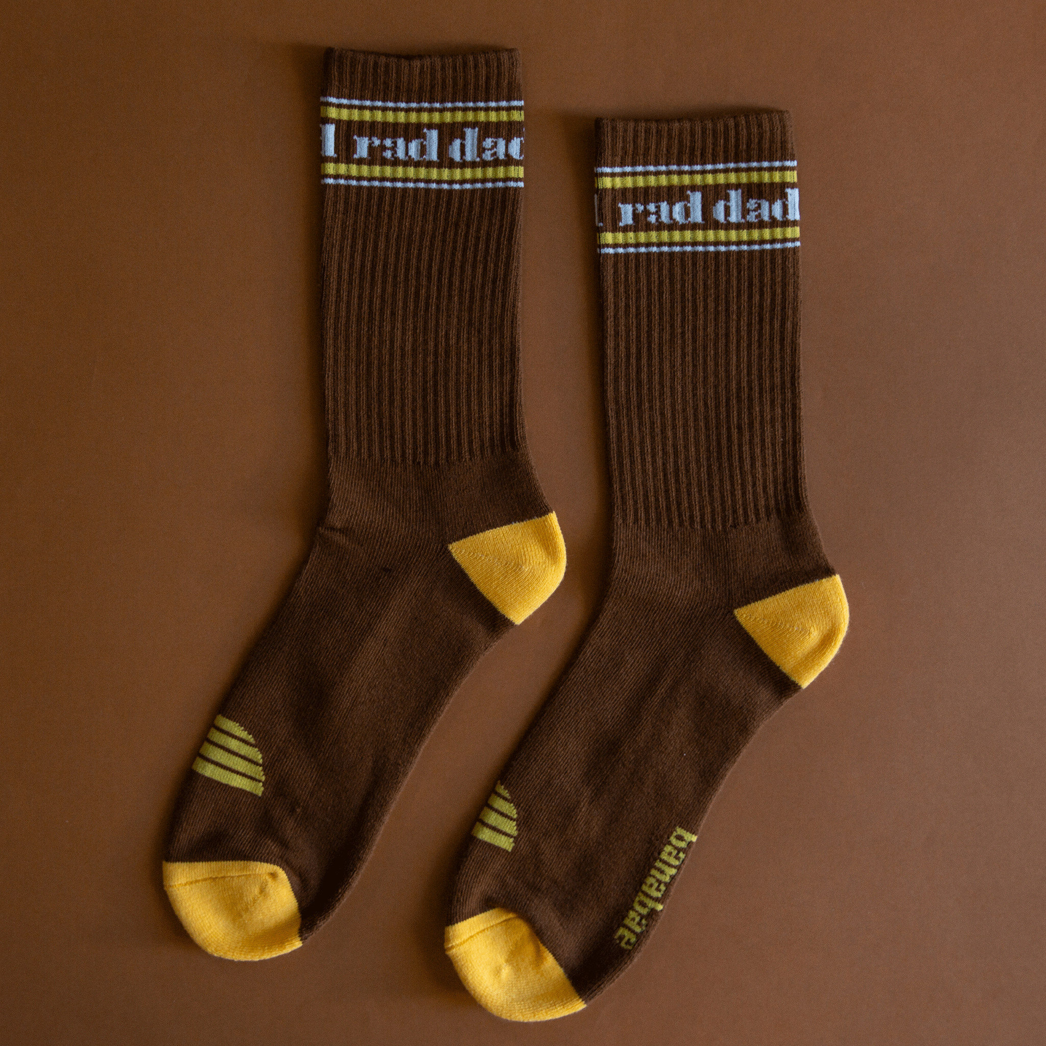 On a brown background is a pair of brown and yellow socks with white text at the top that reads, &quot;rad dad&quot;.