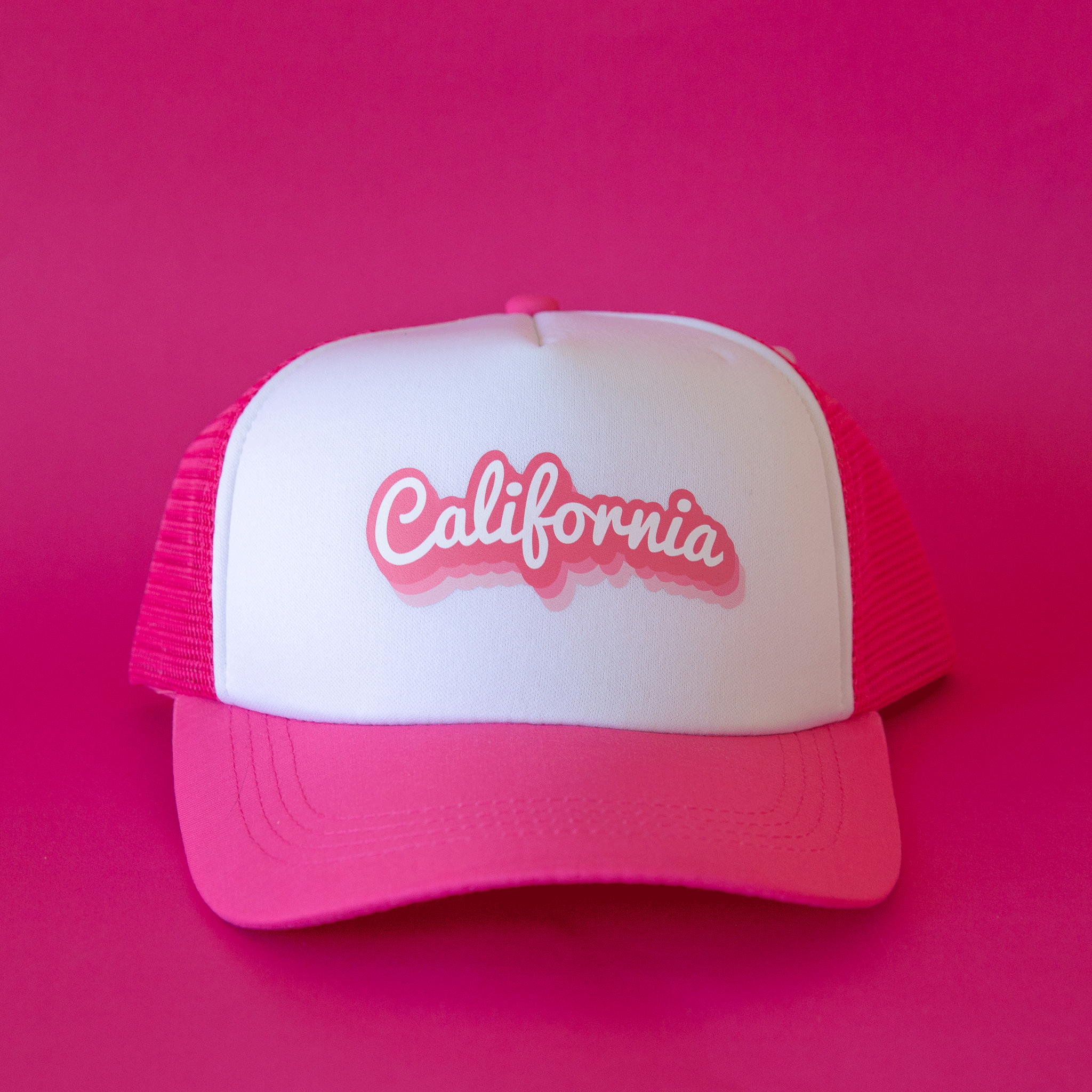 On a hot pink background is a white and hot pink trucker hat with a mesh backing and text across the front that reads, "California".