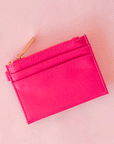 On a pink background is a hot pink card case with a gold zipper and details.