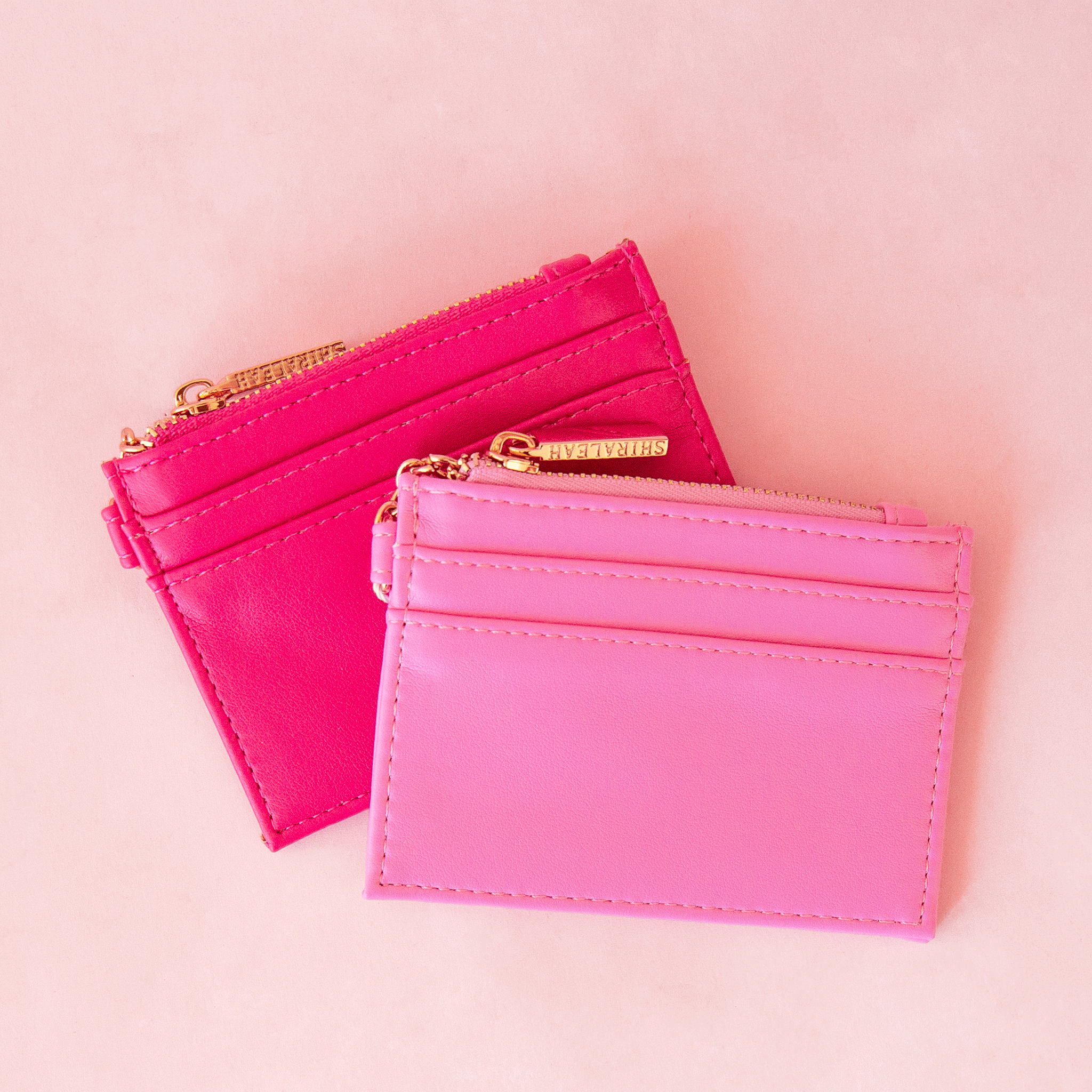 On a pink background is a bubble gum pink card case next to a hot pink version with a gold zipper and details. 