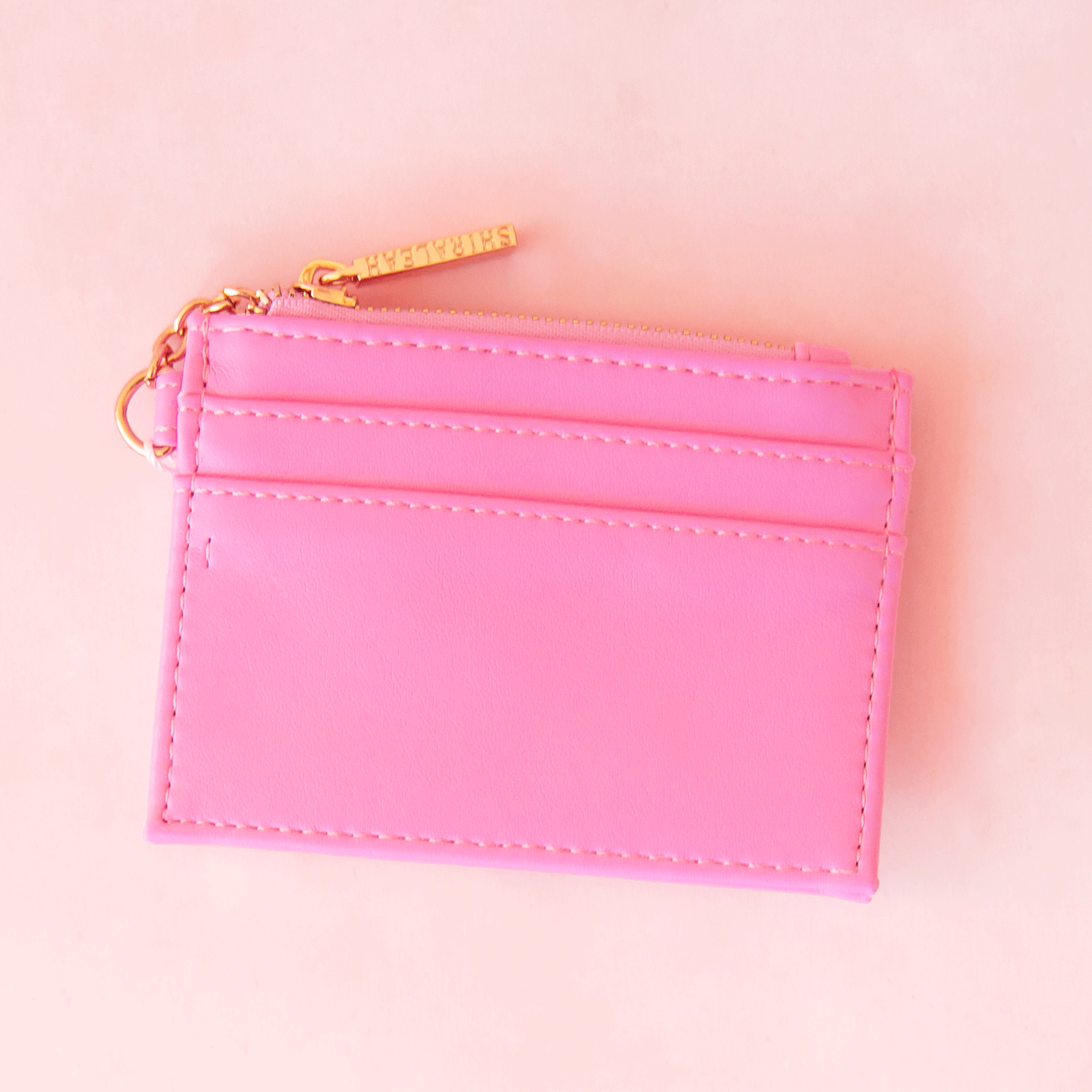 On a pink background is a bubble gum pink card case with a gold zipper and details. 