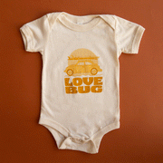 On a burnt orange background is an ivory short sleeve children's onesie with a yellow VW bug with a surfboard graphic with orange letters underneath that reads, "Love Bug". 