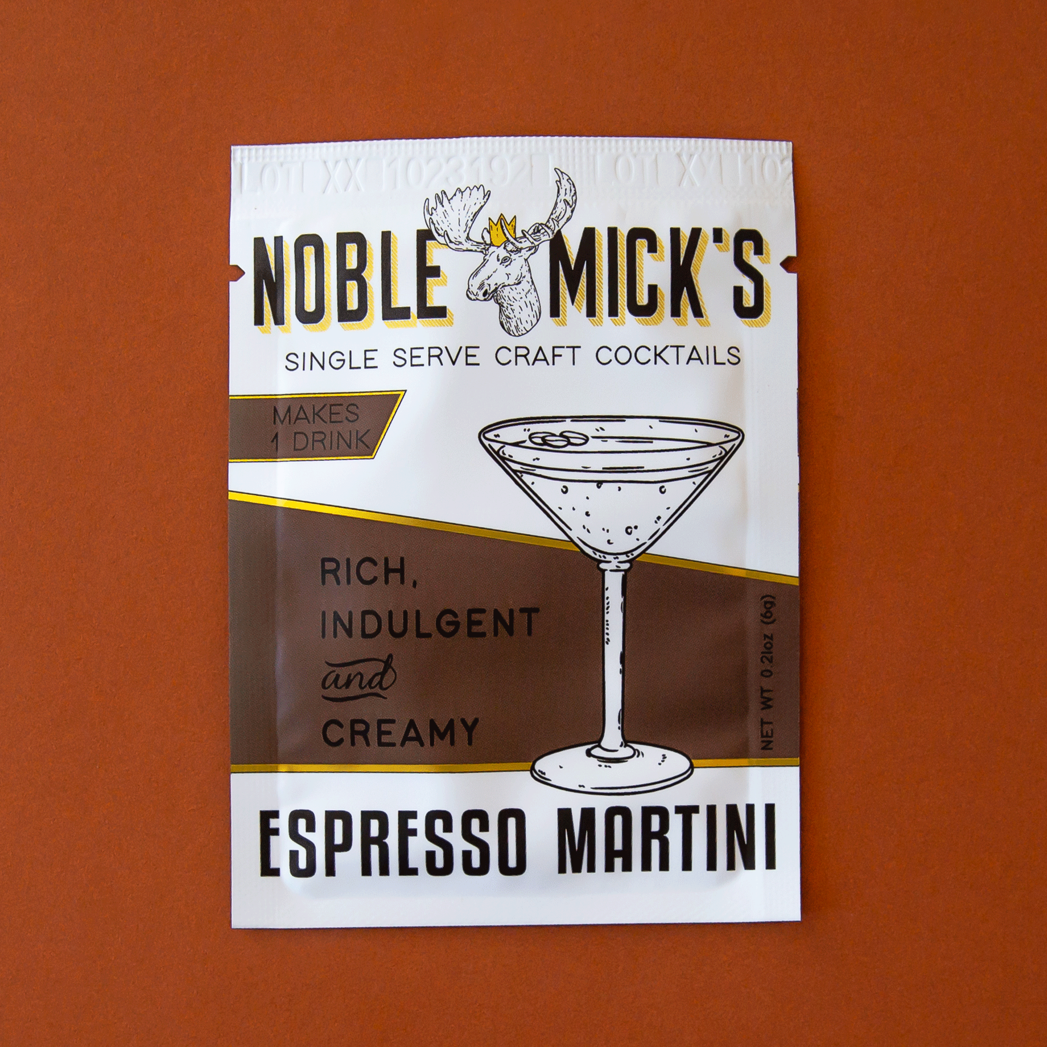 On a burnt orange background is a white and brown cocktail mix packet for espresso martinis with a graphic of a martini glass and a moose at the top that reads, &quot;Noble Mick&quot;s Single Serve Craft Cocktails&quot;.