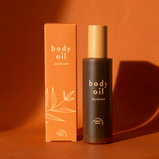 On an orange background is a bottle of body oil with a tan cap and an orange box packaging along with text on the front that reads, "body oil daydream".