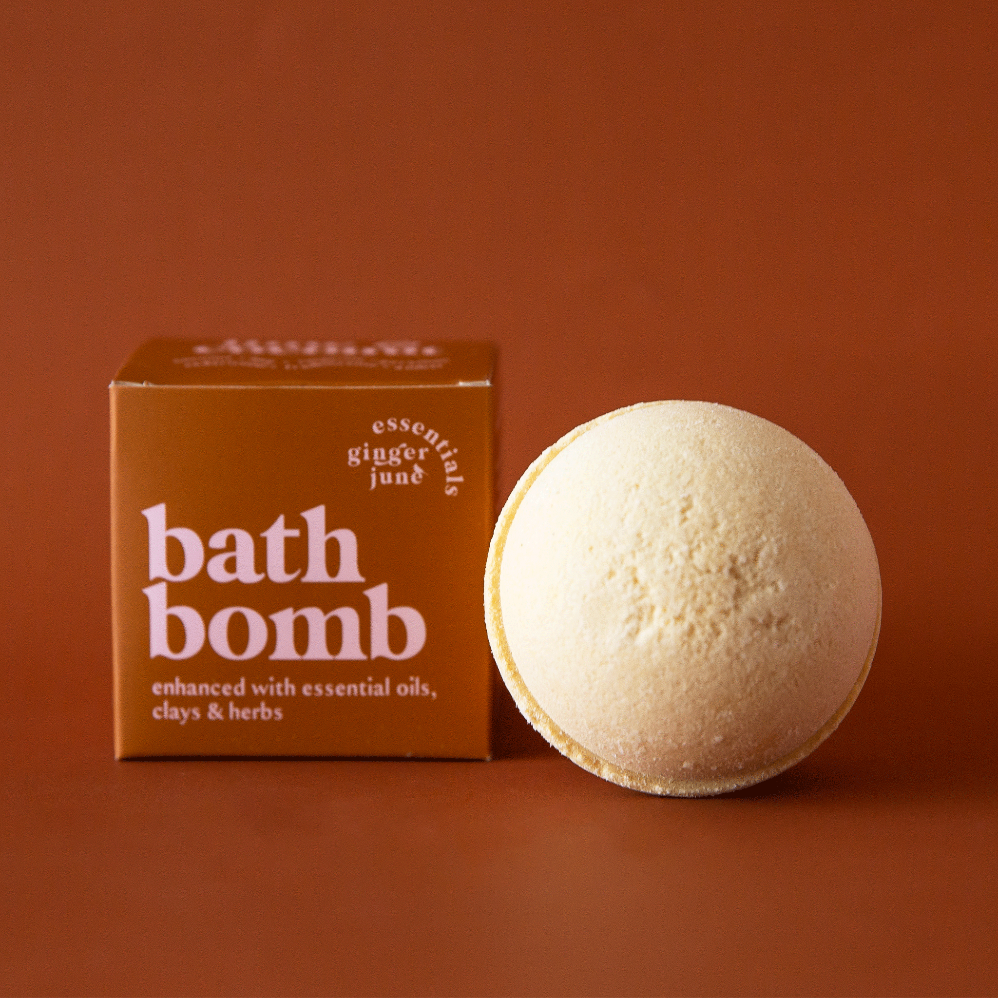 On a brown background is a warm brown box with a tan bath bomb next to it. The text on the box reads, "bath bomb enhanced with essential oils, clays and herbs".