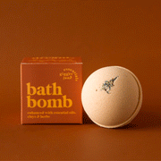On a burnt orange background is a round bath bomb with an orange hue and a orange box with yellow text that reads, "bath bomb enhanced with essential oils, clays & herbs".