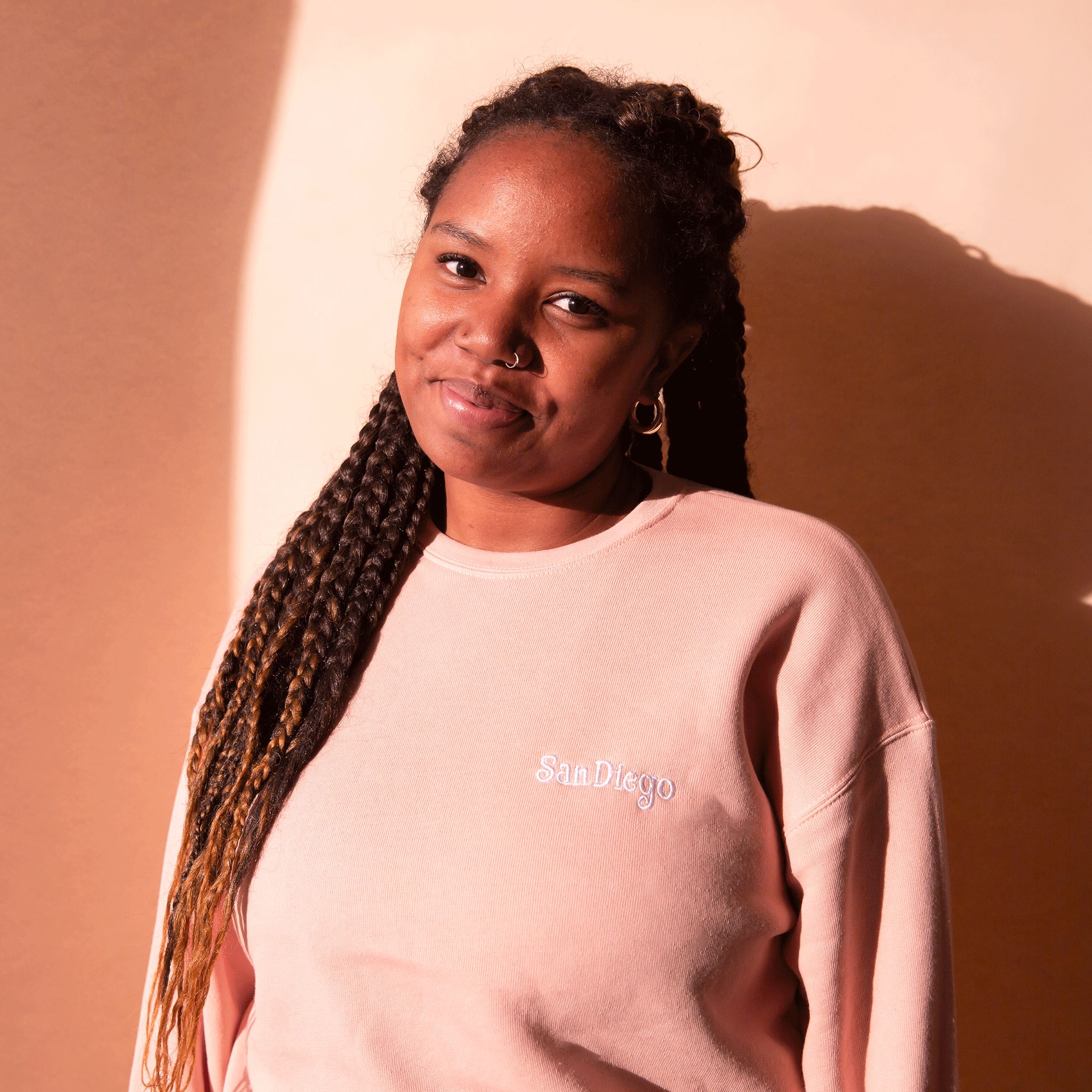 On a peach background is a peachy pink pull over sweatshirt with a white embroidered &quot;San Diego&quot; on the front.
