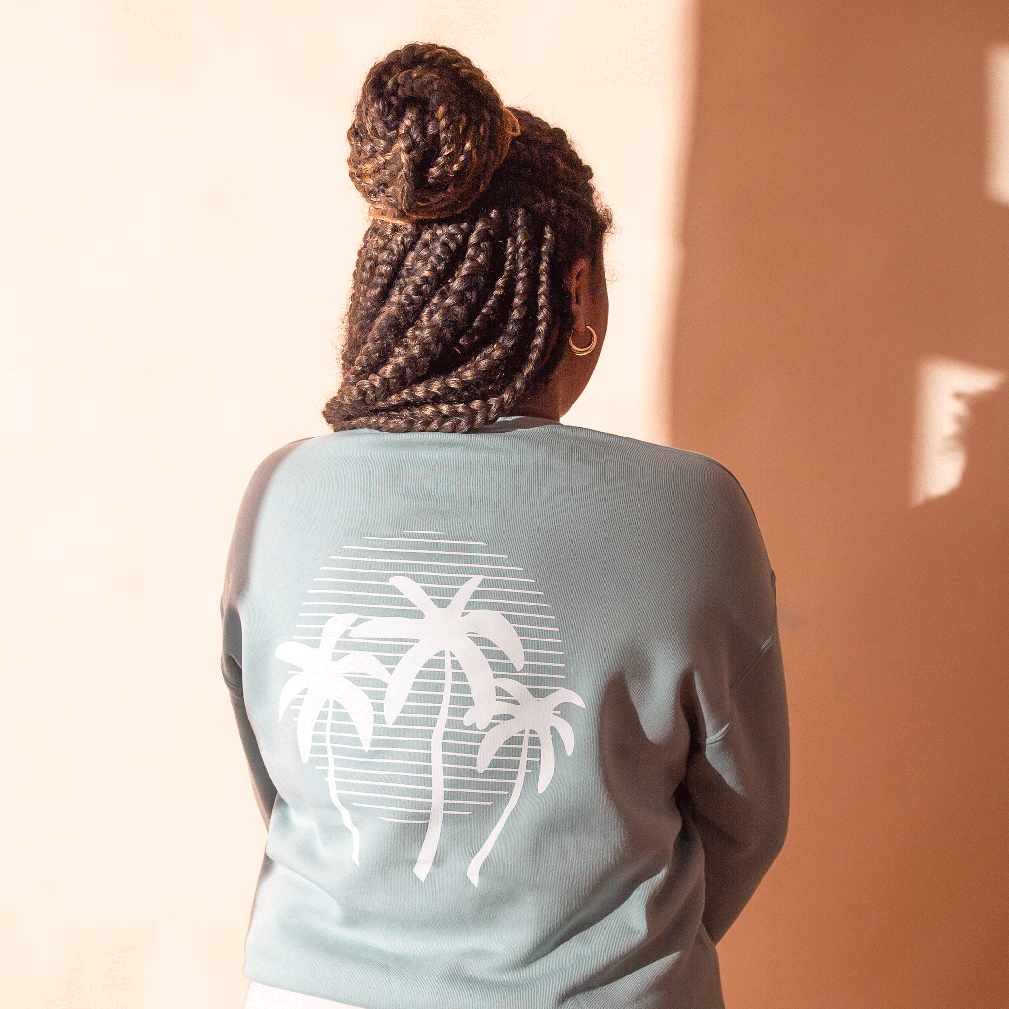 On a peach background is a light blue crewneck sweatshirt with a white palm tree sunset graphic on the back. 