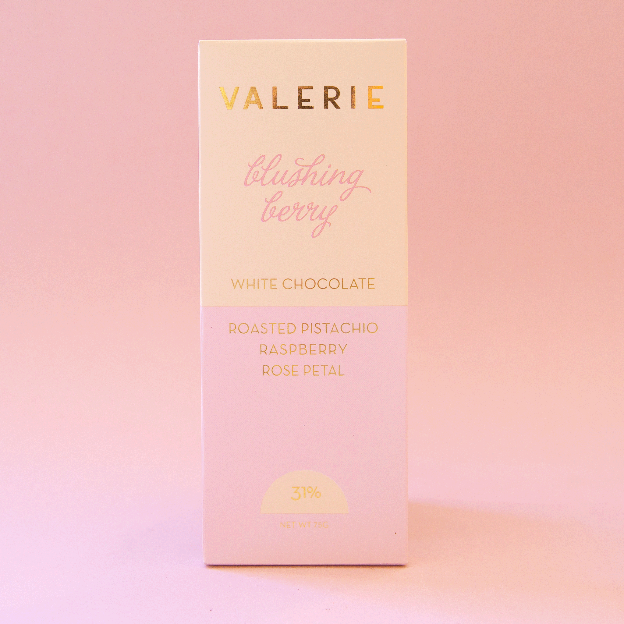 On a pink background is a light pink and ivory packaged white chocolate bar that reads, "Valerie blushing berry White Chocolate Roasted Pistachio, Raspberry, Rose Petal".