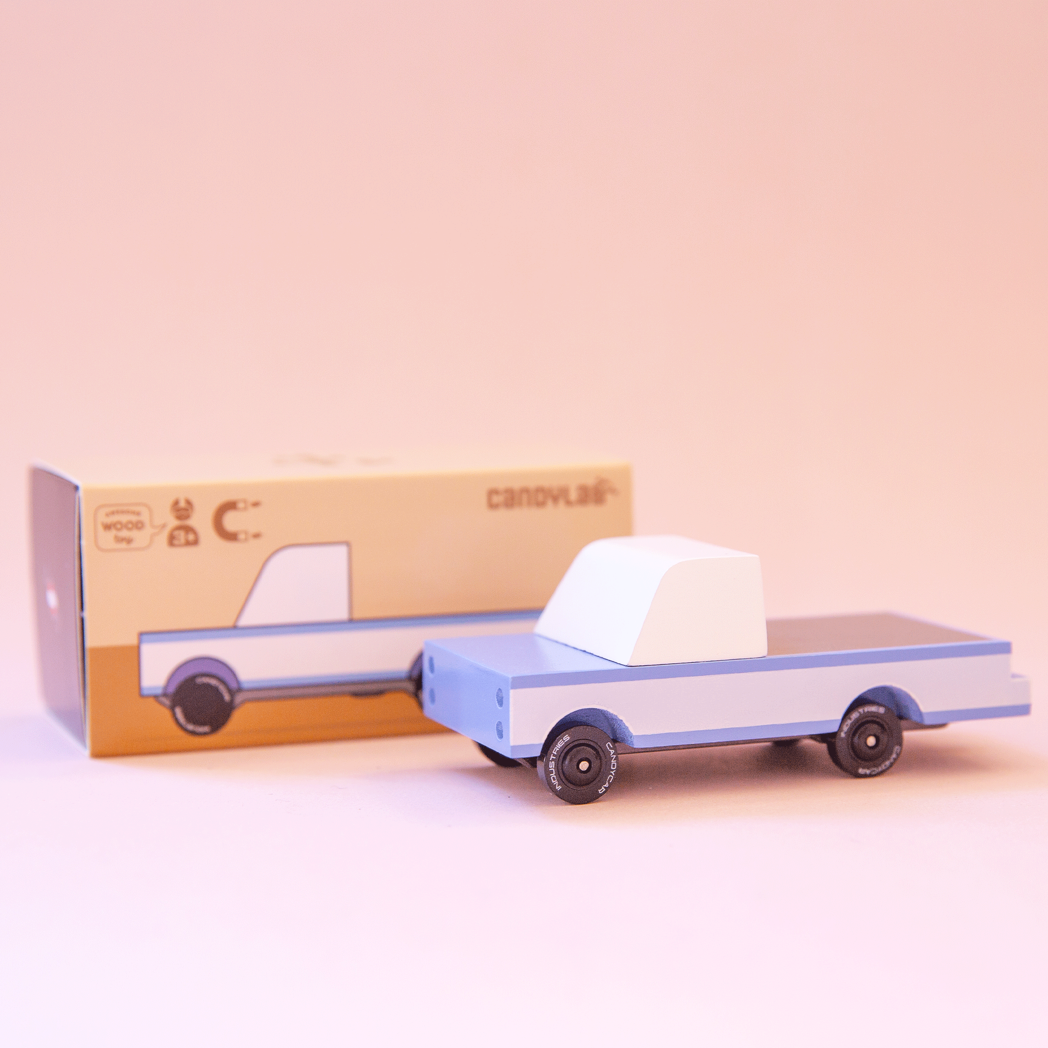 A blue wooden toy pickup truck on a peachy background in front of the box it comes in. 