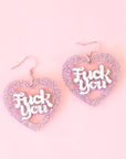 A pair of silver glitter heart shaped earrings with white text in the center that reads, "Fuck You". 
