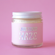 On a pink background is a 4oz glass jar candle with a light pink label that reads, "Lavender Haze" in a holographic wavy font.