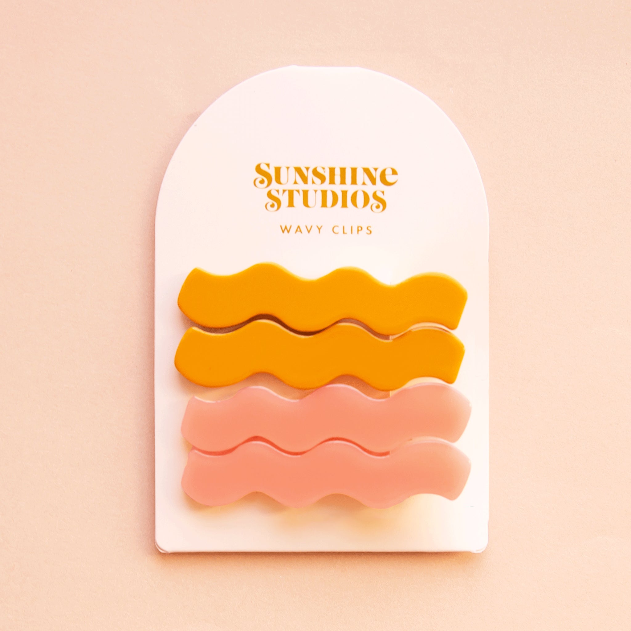 On a pink background two sets of wavy hair clips in a mango orange shade and a light pink shade clipped on a white arched piece of cardboard packaging with orange text at the top that reads, "Sunshine Studios".