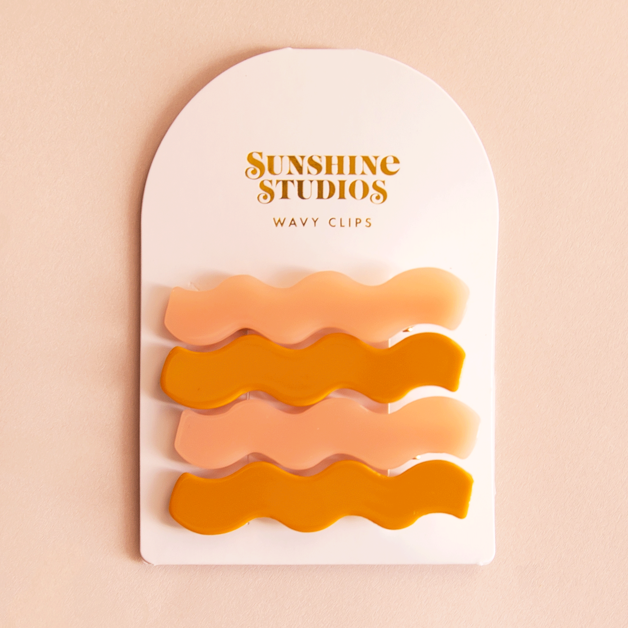 On a pink background two sets of wavy hair clips in a mango orange shade and a light pink shade clipped on a white arched piece of cardboard packaging with orange text at the top that reads, &quot;Sunshine Studios&quot;.