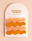 On a pink background two sets of wavy hair clips in a mango orange shade and a light pink shade clipped on a white arched piece of cardboard packaging with orange text at the top that reads, "Sunshine Studios".