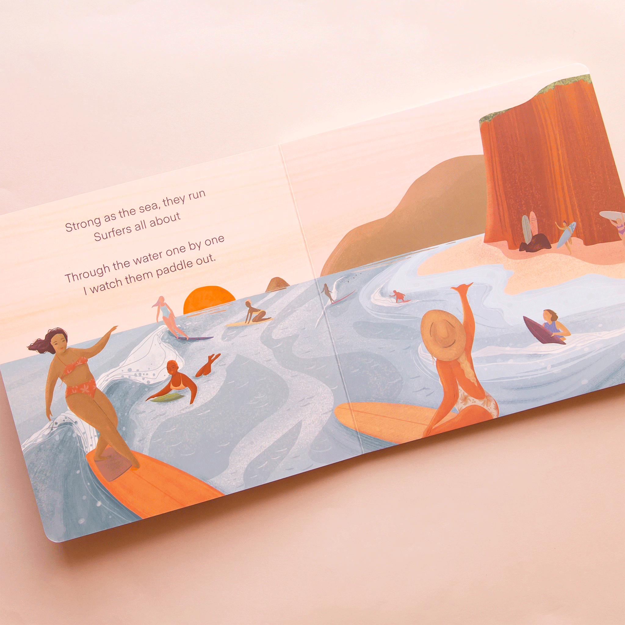 On a pink background is the book open to a page with illustrations of a beach and the ocean along with women in the water surfing and text that reads, "Strong as the sea they run Surfers all about Through the water one by one I watch them paddle out". 