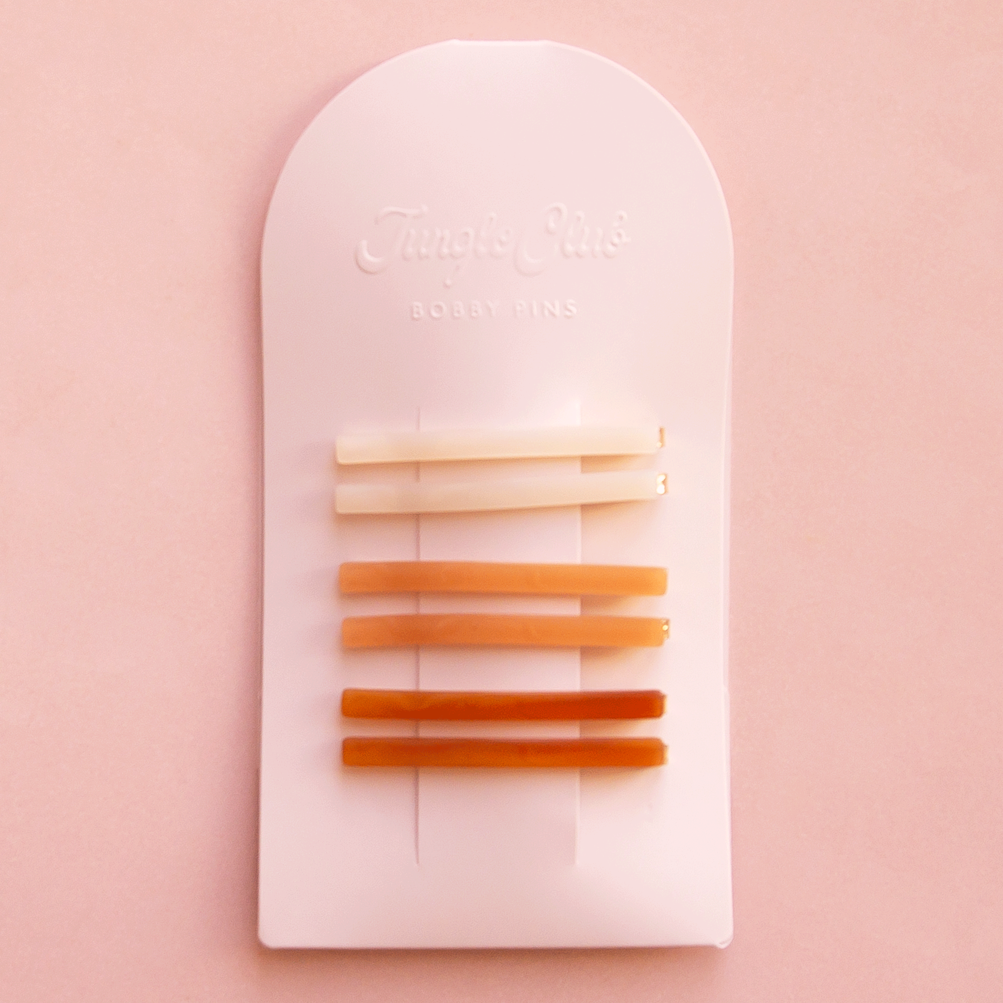 On a pink background is an arched packaging with embossed text at the top that reads, &quot;Jungle Club Bobby Pins&quot; along with six thin hair clips in three colors from an ivory shade to orange to a honey rust color. 