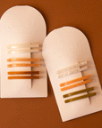 On a burnt orange background is both of the available color ways of the acetate bobby pin sets on arched packaging. 