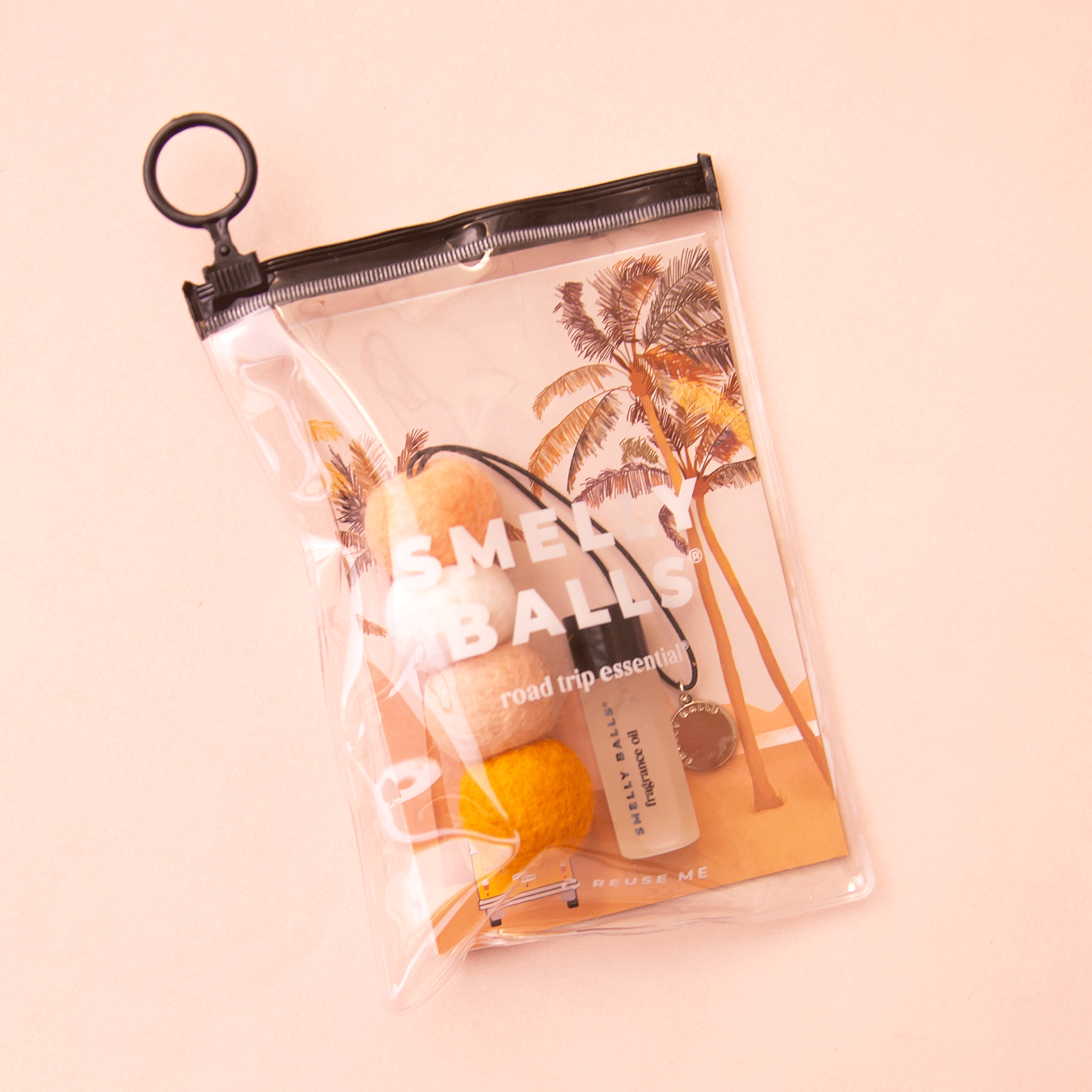 On a peachy background is a clear pouch with a black zipper filled with four small wool balls made into an air freshener with a small bottle of fragrance oil next to it.