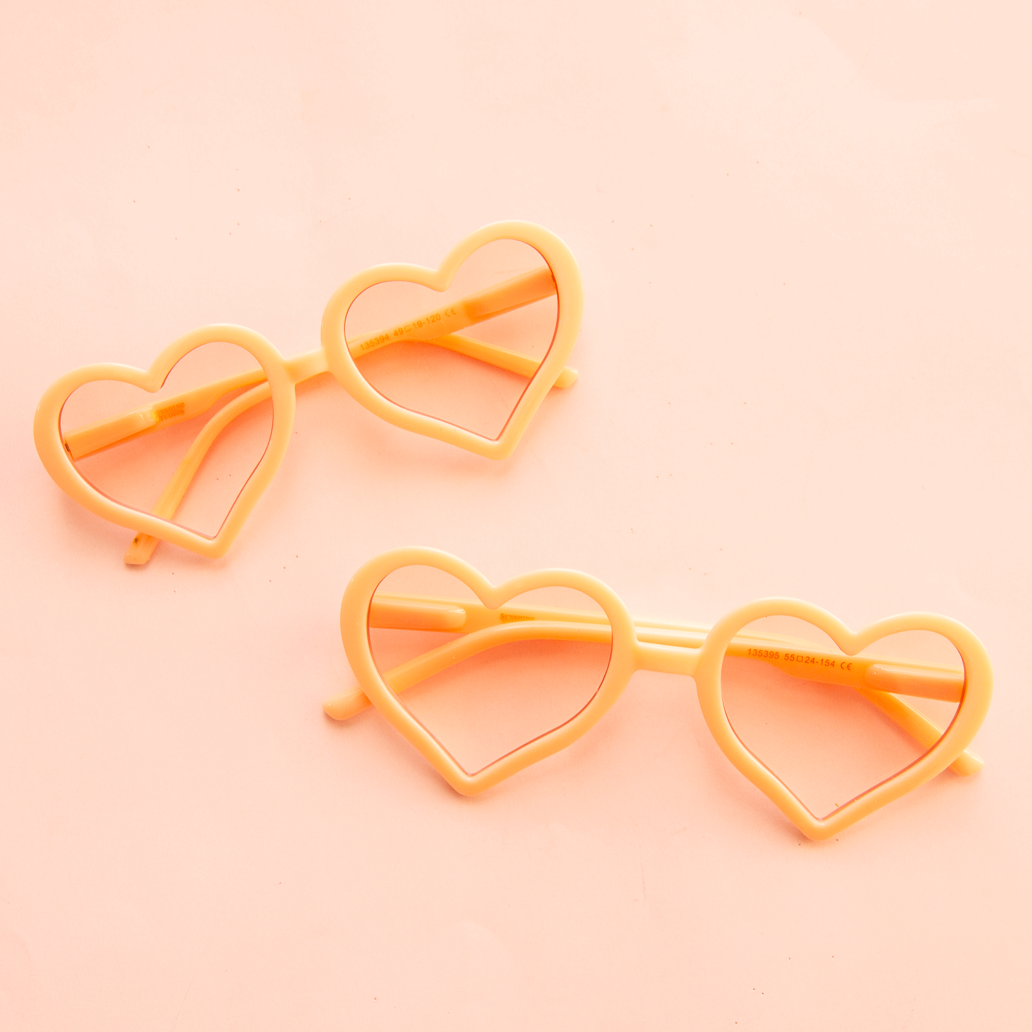 On a neutral background is two pairs of heart shaped glasses in an apricot shade shown here in two sizes, one for kids and one for adults. 
