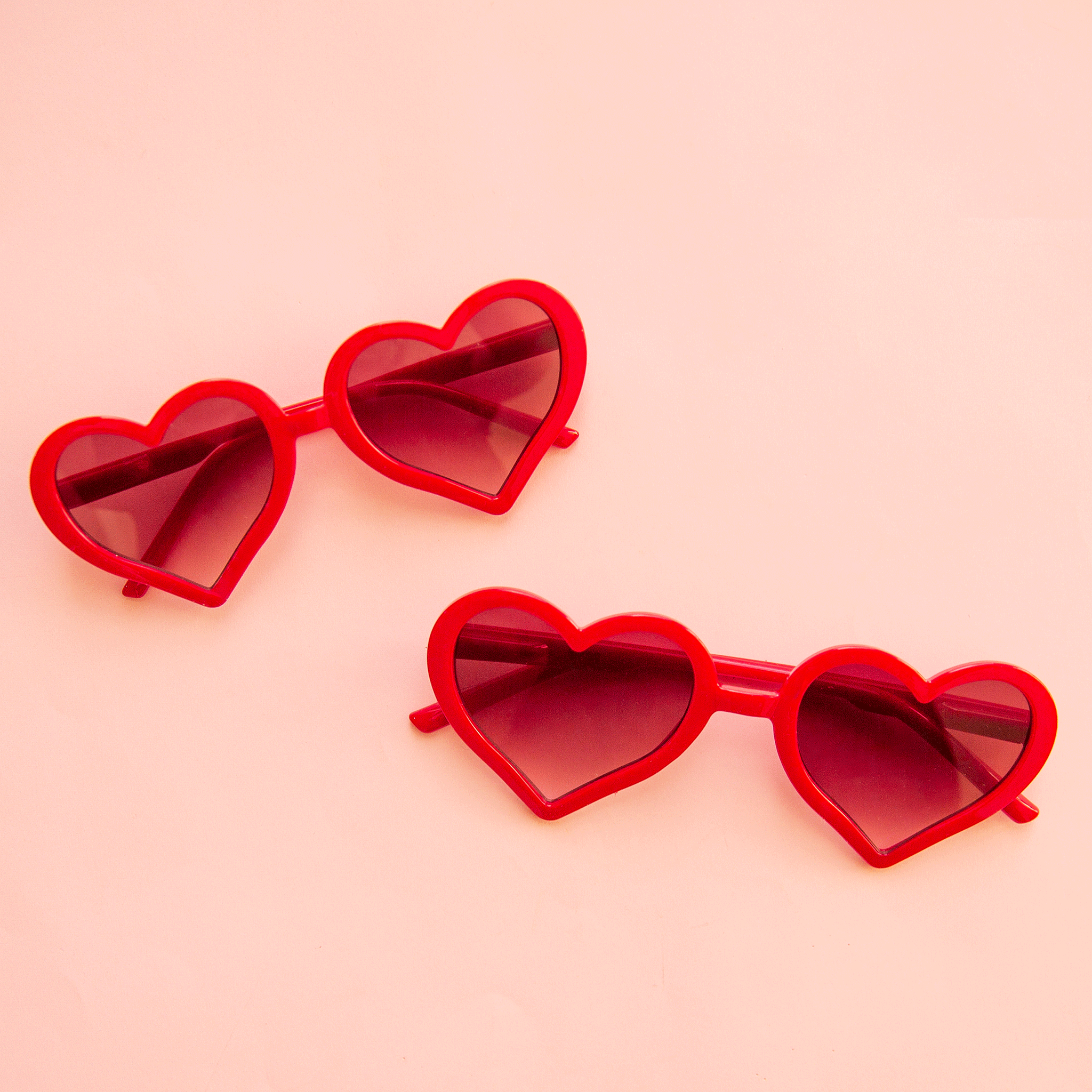 On a pink background is a red pair of heart shaped sunglasses in two different sizes, one for kids and one for adults.
