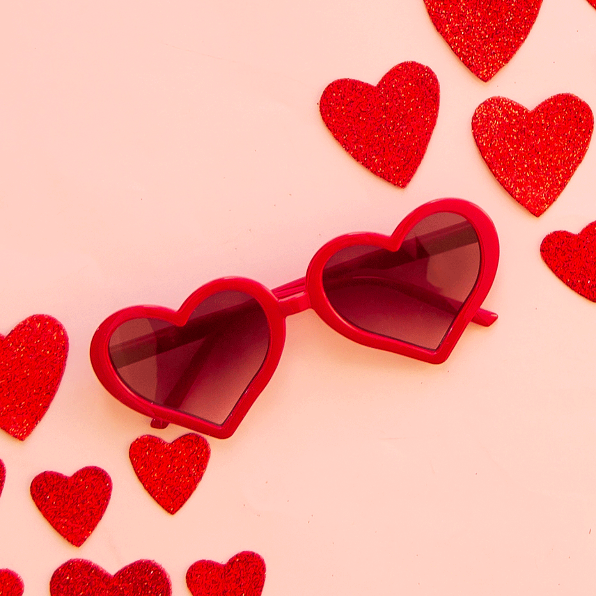 On a pink background is a red pair of heart shaped sunglasses. 