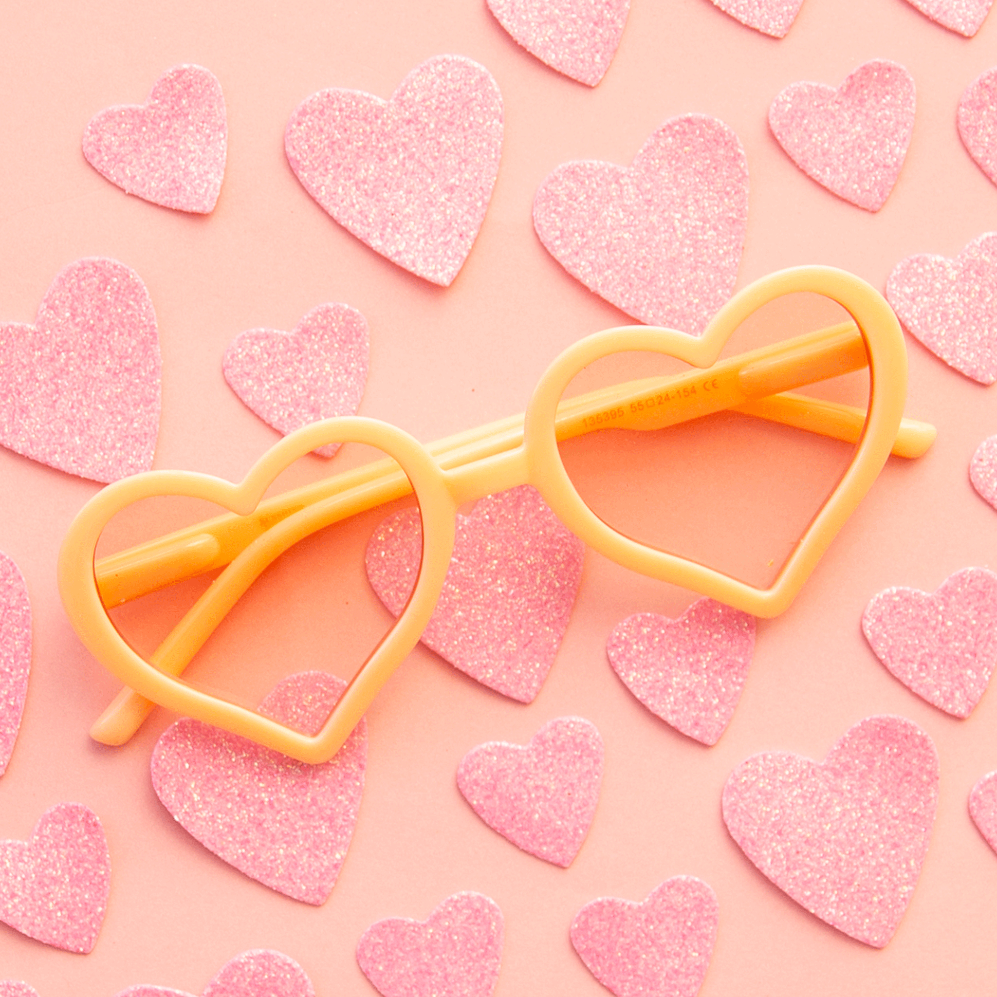 On a pink background with pink glitter hearts is a pair of apricot colored heart shaped sunglasses. 