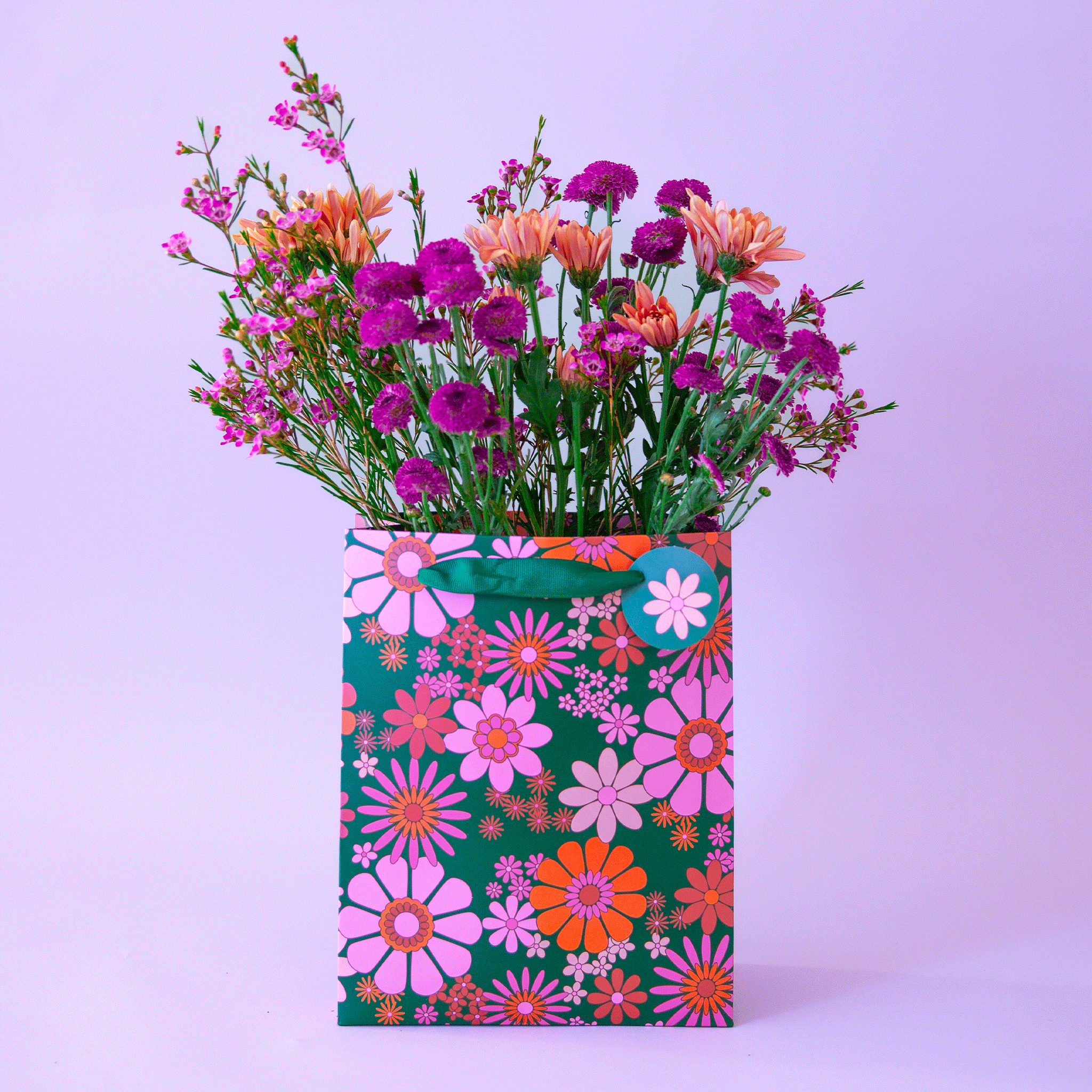 On a purple background is a green gift bag with a red, pink and purple floral print and filled here with flowers that are not included with purchase. 