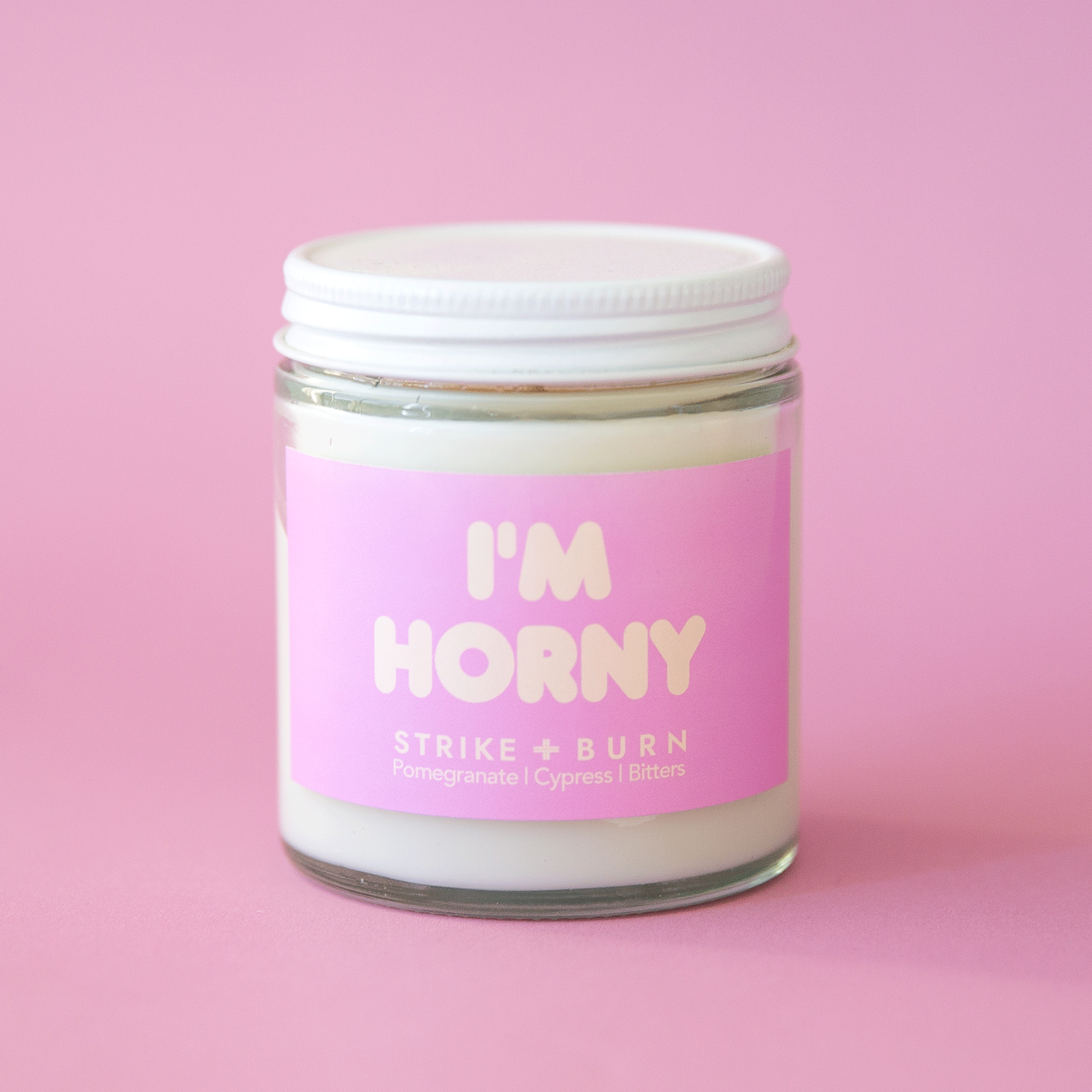A glass jar with a white wax candle featuring a orchid purple colored label along with text that reads, "I'm Horny" in cream letters.