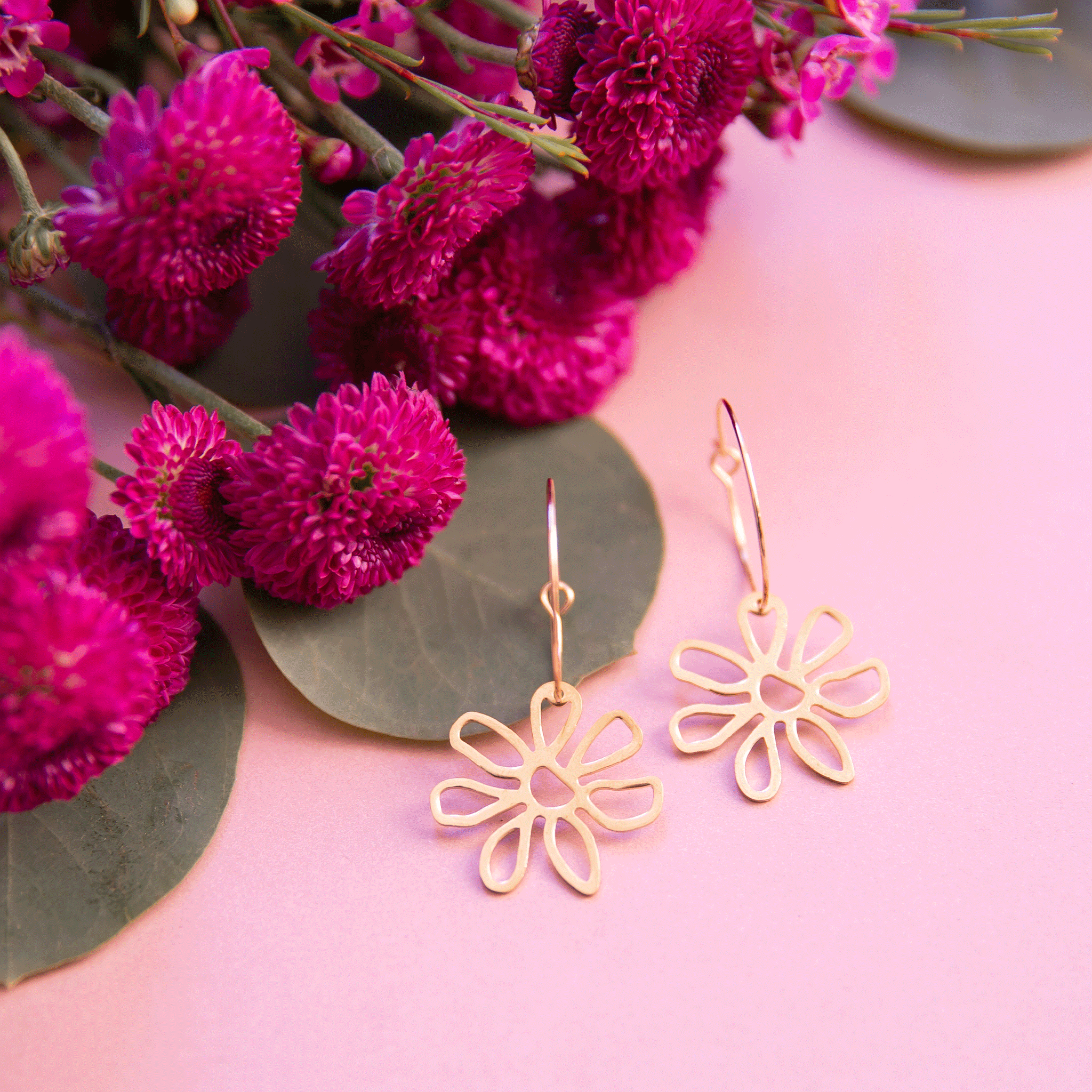 On a pink background is a pair of gold hoop earrings with gold flower outlines hanging from the bottom of each hoop.