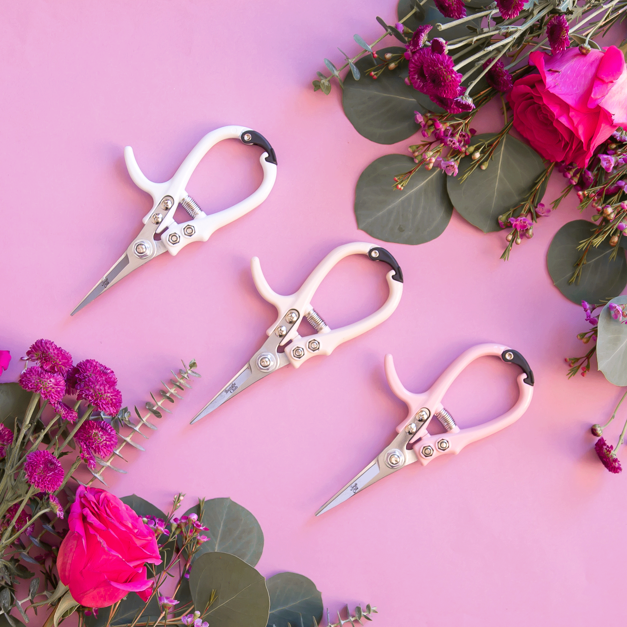 On a pink background is all three color ways of the pruning shears next to bouquets of flowers. 