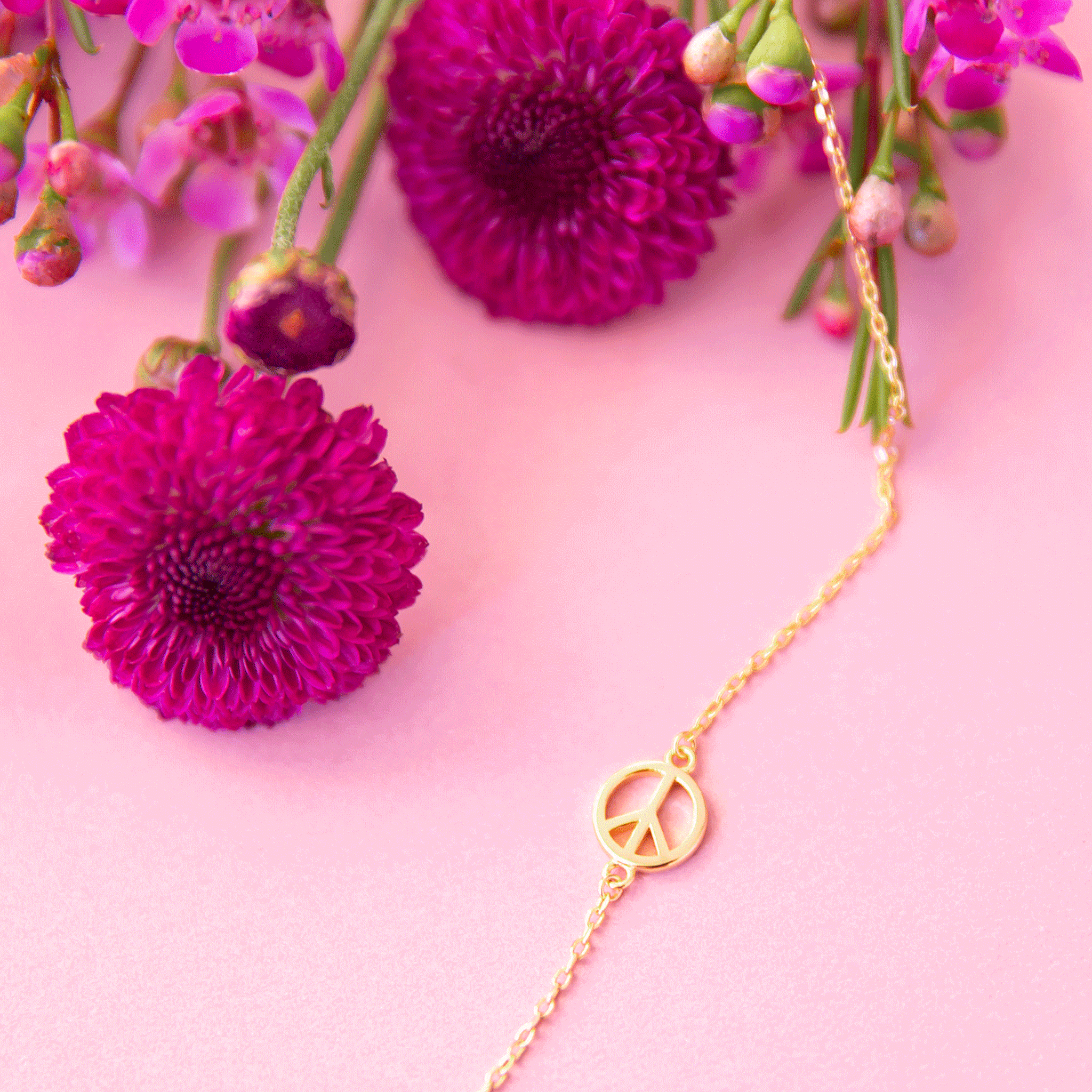 On a pink background is a gold chain necklace with a gold peace sign on the necklace.