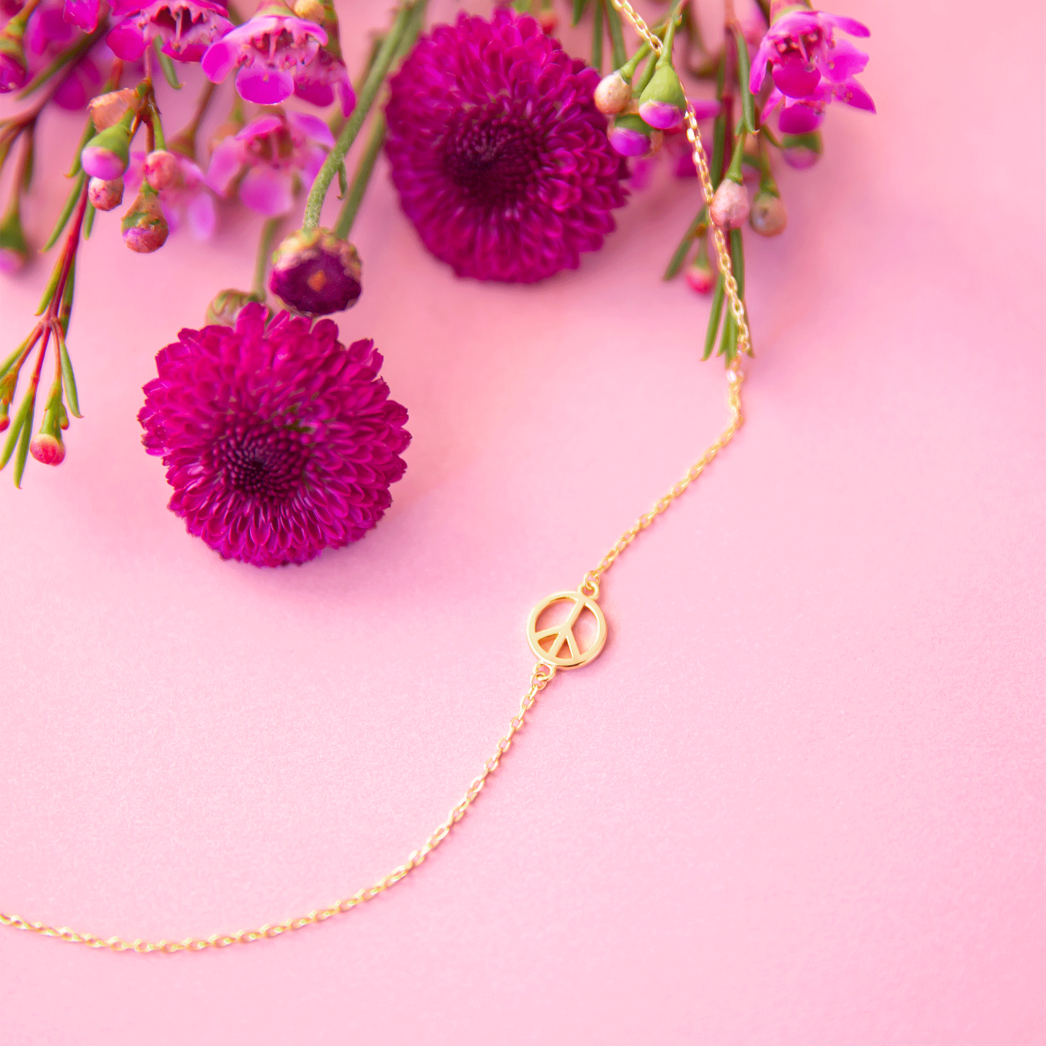 On a pink background is a gold chain necklace with a gold peace sign on the necklace. 